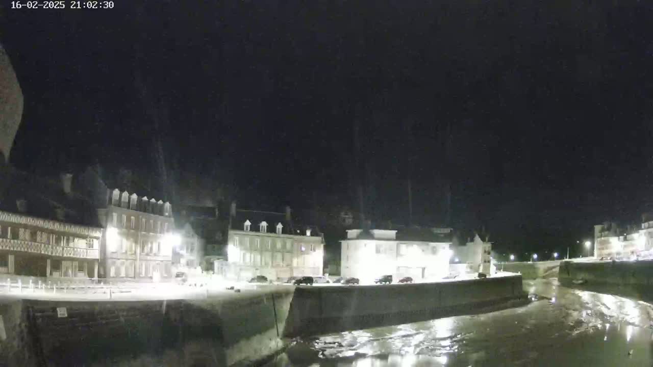 At night, several buildings are illuminated along a waterway with a low tide, under a dark, clear sky.