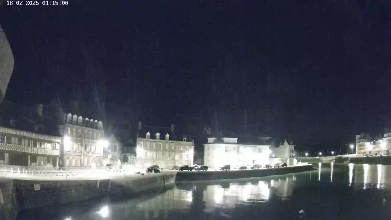 A nighttime view of a calm canal reflecting illuminated buildings along its banks under a clear, dark sky.