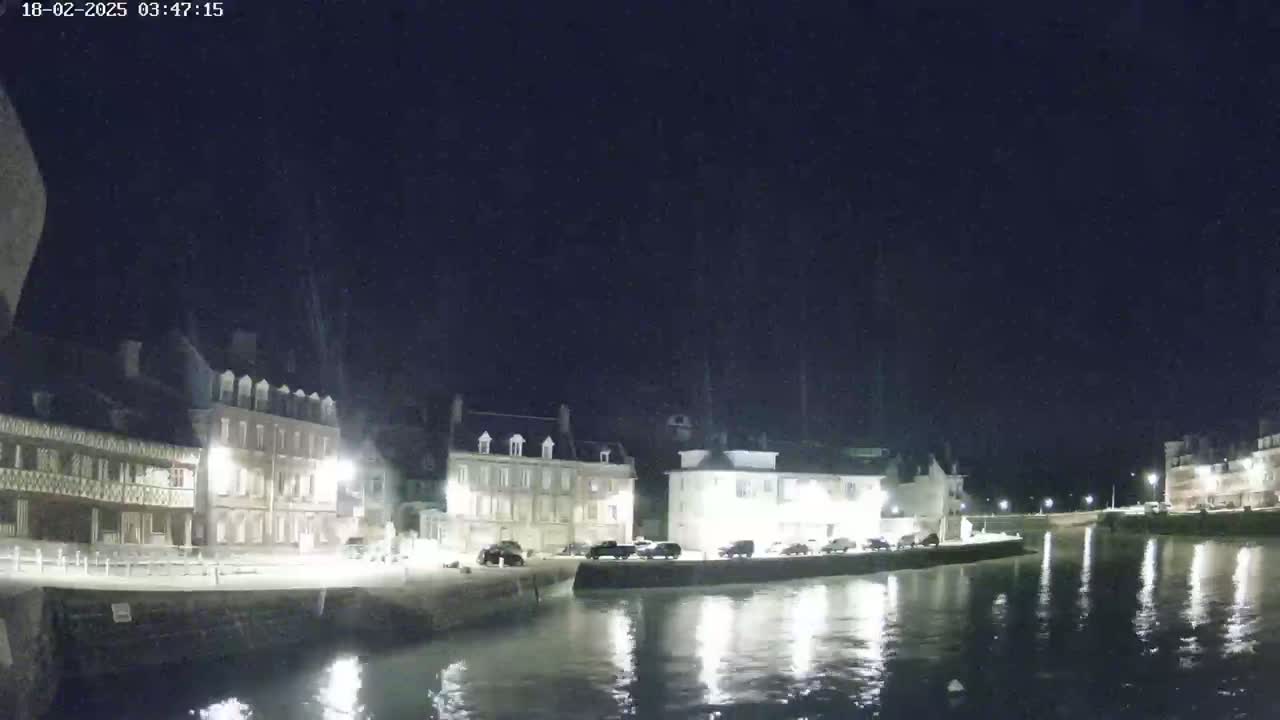 A nighttime view of a calm waterway reflecting lights from buildings lining the waterfront under a clear, dark sky.