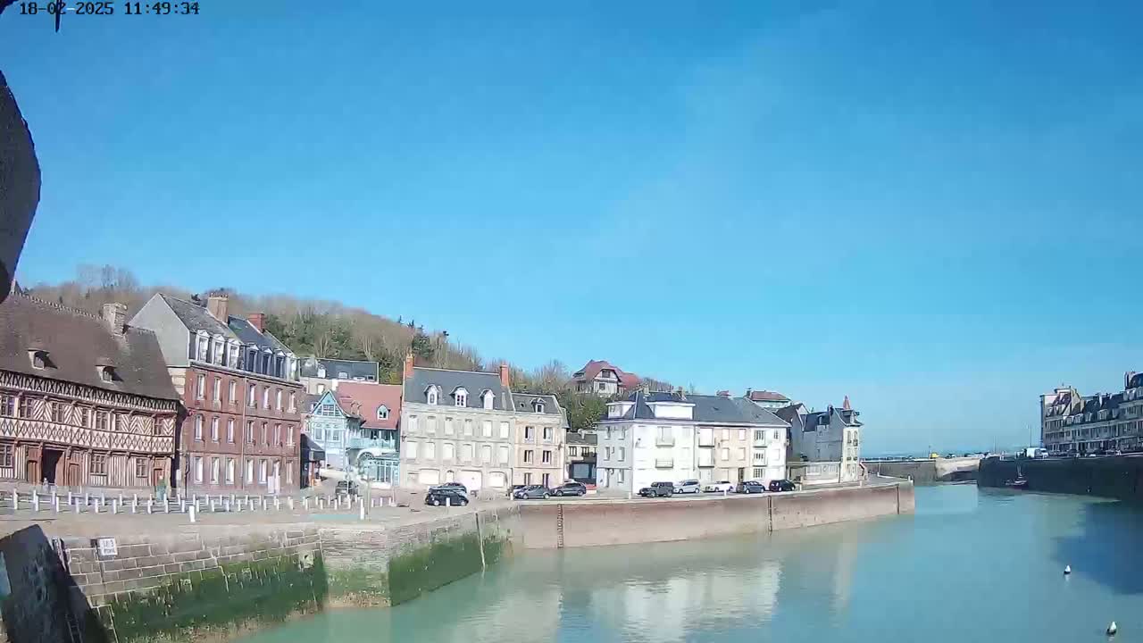 A waterfront town with multi-story buildings lines a calm canal under a clear blue sky.