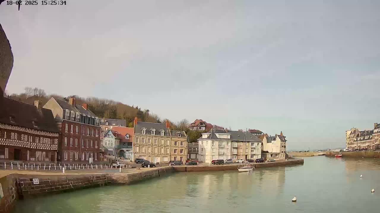 A calm harbor is seen with several multi-story buildings lining the waterfront under a mostly cloudy sky.
