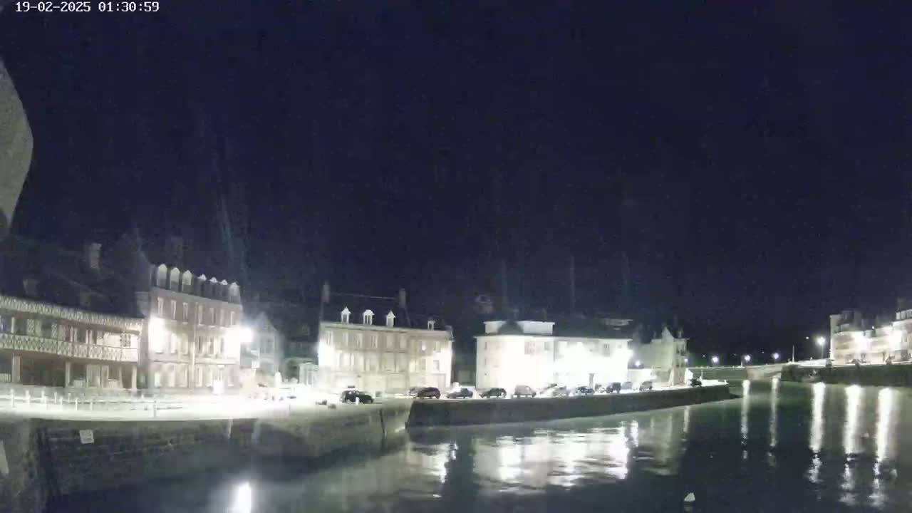 A nighttime view of a waterfront town with several buildings illuminated along a calm body of water under a clear, dark sky.