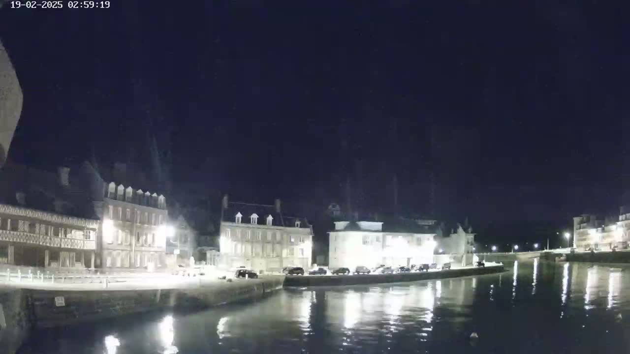 At night, several multi-story buildings line a calm waterway reflecting lights, under a clear, dark sky.