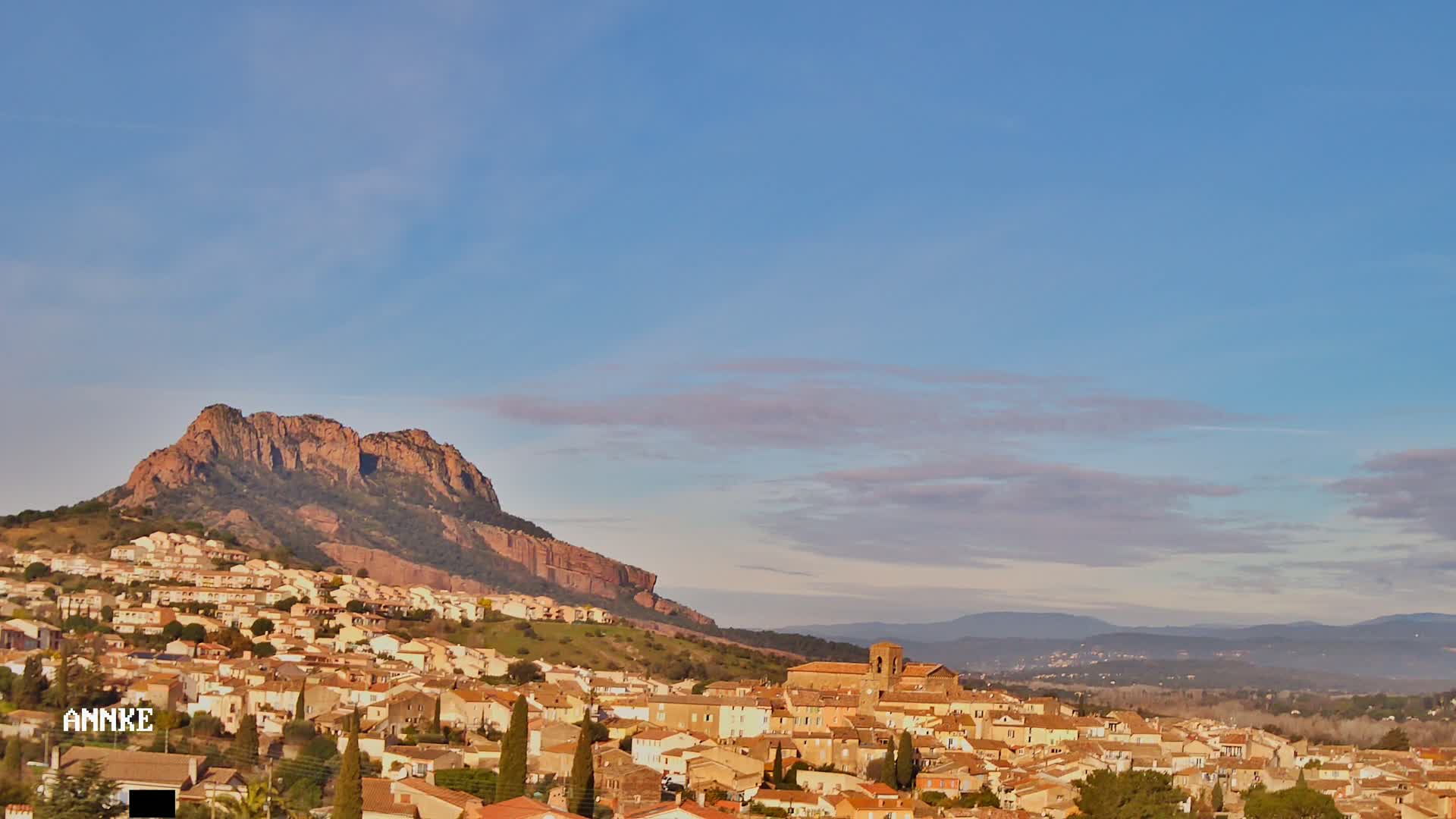 A small town with tan and brown buildings is nestled at the base of a large, reddish-brown rock formation under a mostly clear, light-blue sky.