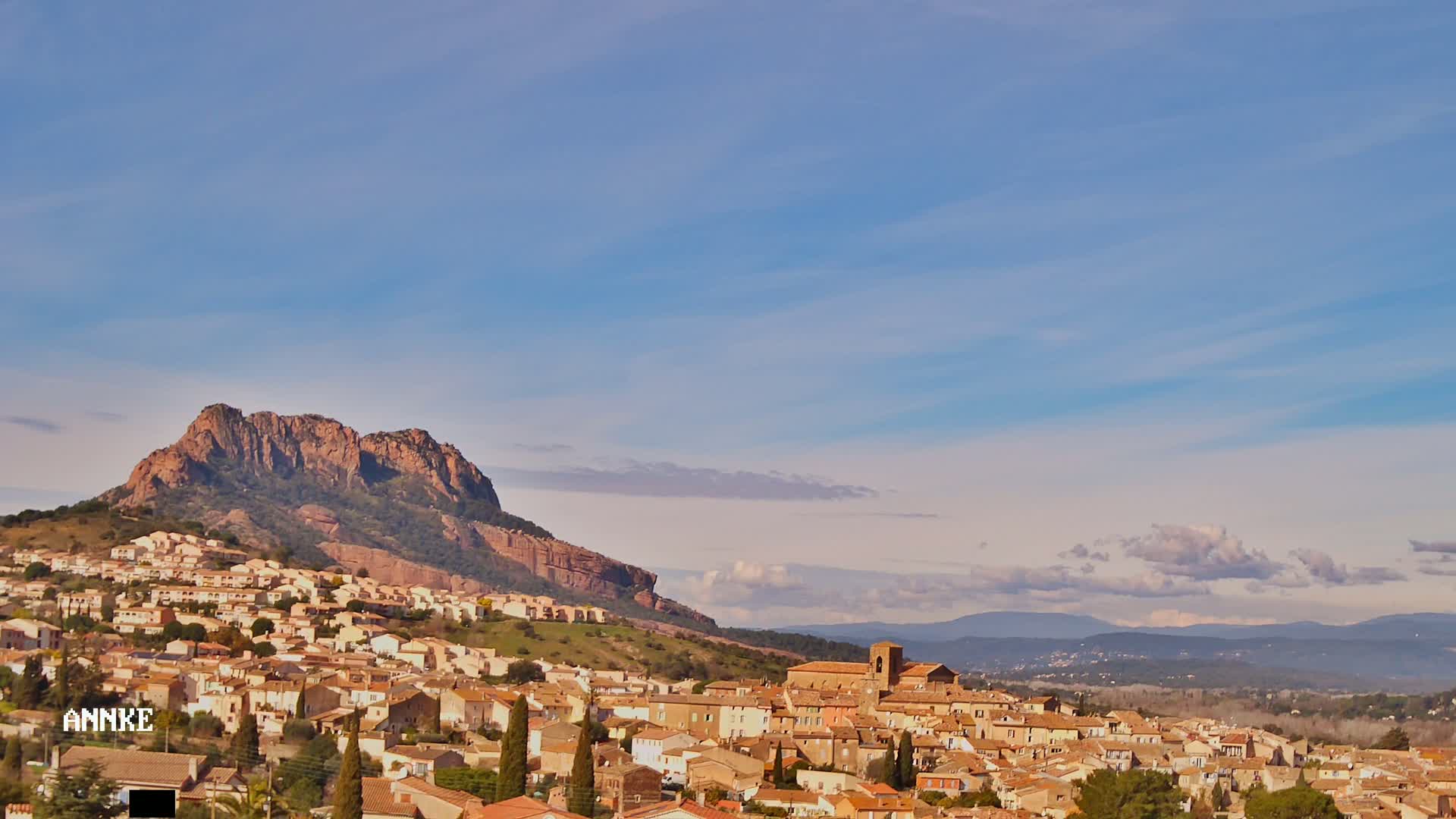 A small town of tan buildings nestles at the base of a large, reddish-brown mountain under a mostly clear, light-blue sky with a few wispy clouds.