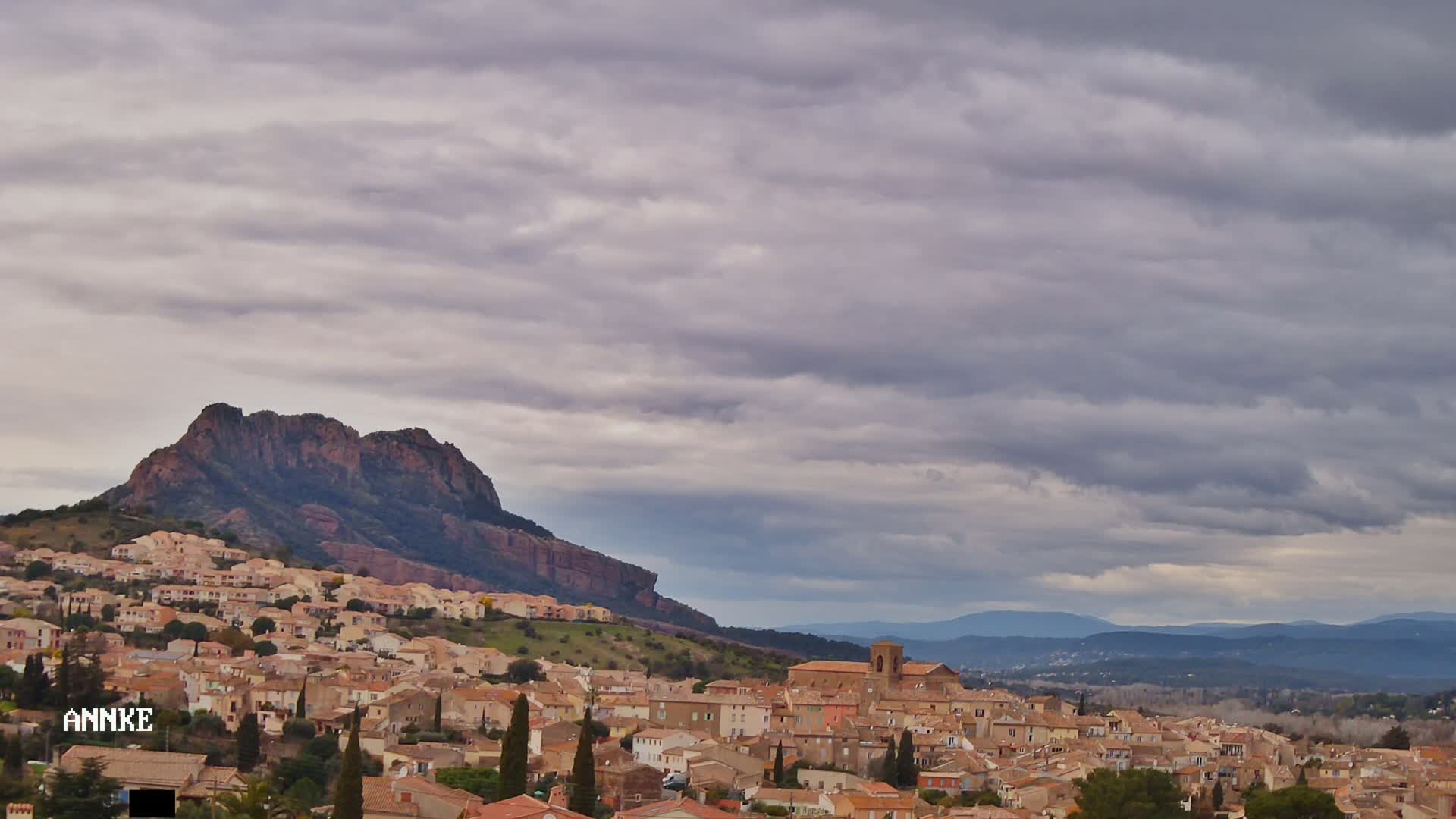 A sprawling town of tan and brown buildings nestled on a hillside beneath a large, dark-colored mountain under an overcast sky.