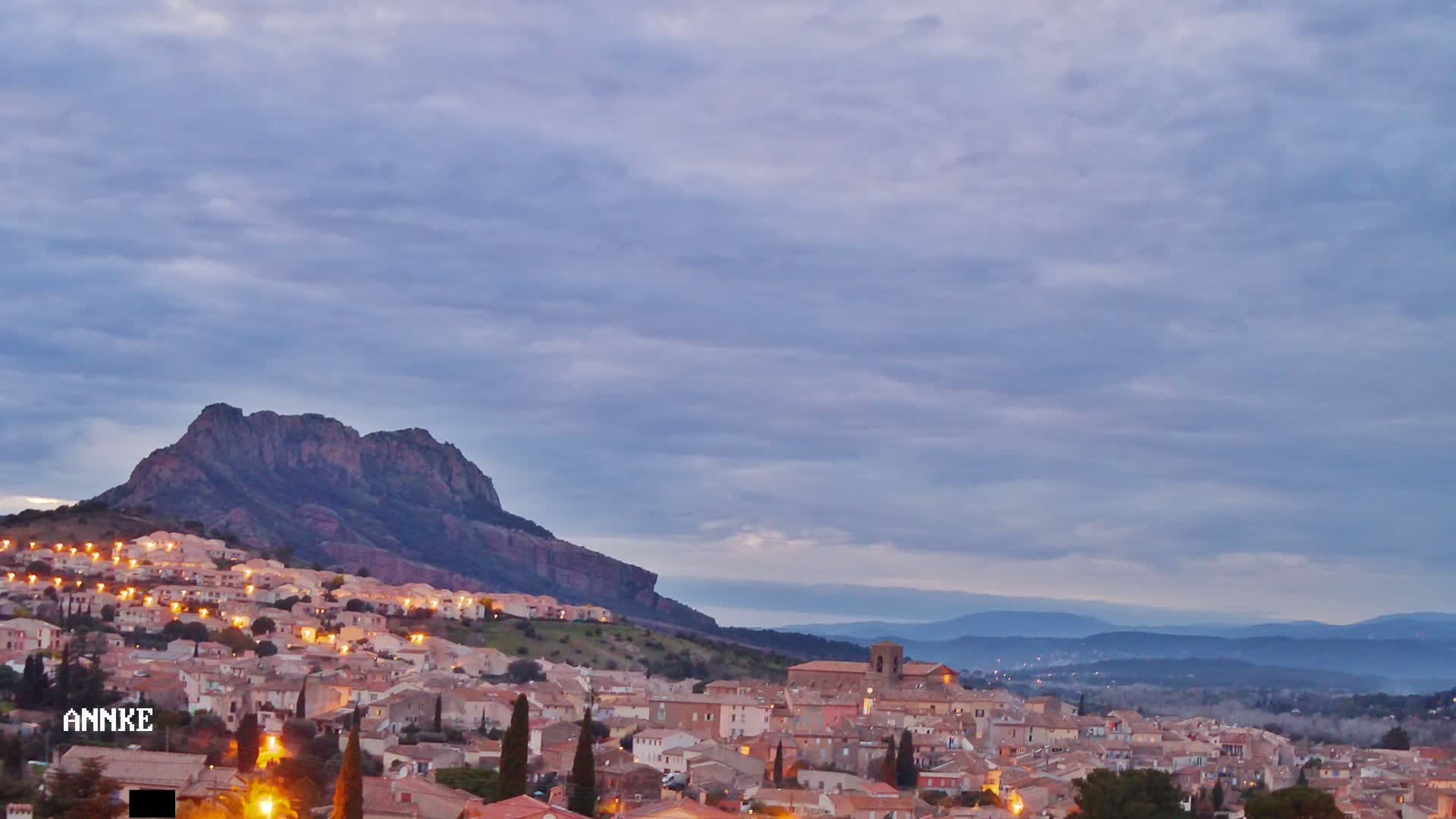 A town with illuminated buildings is nestled at the base of a large, rocky mountain under a mostly cloudy, dusky sky.