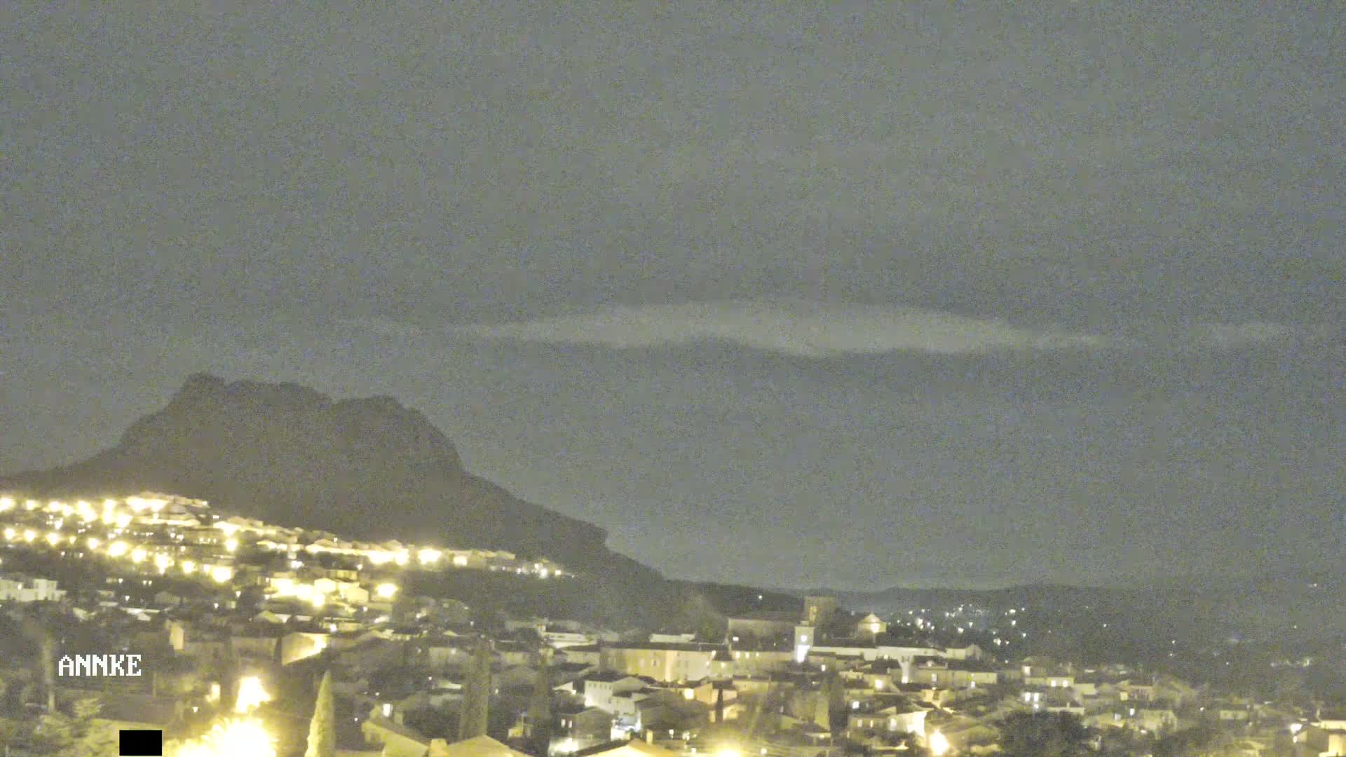 A nighttime view of a town nestled against a large, dark mountain under a mostly cloudy sky.