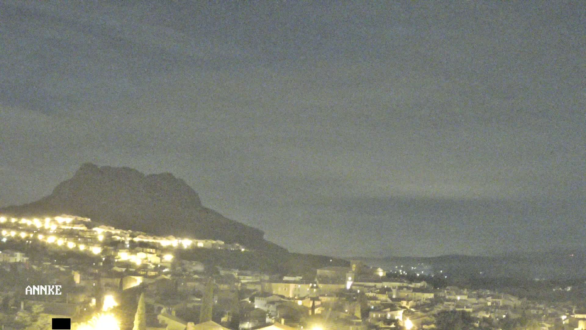 A nighttime view of a town nestled at the base of a dark mountain under a cloudy sky.