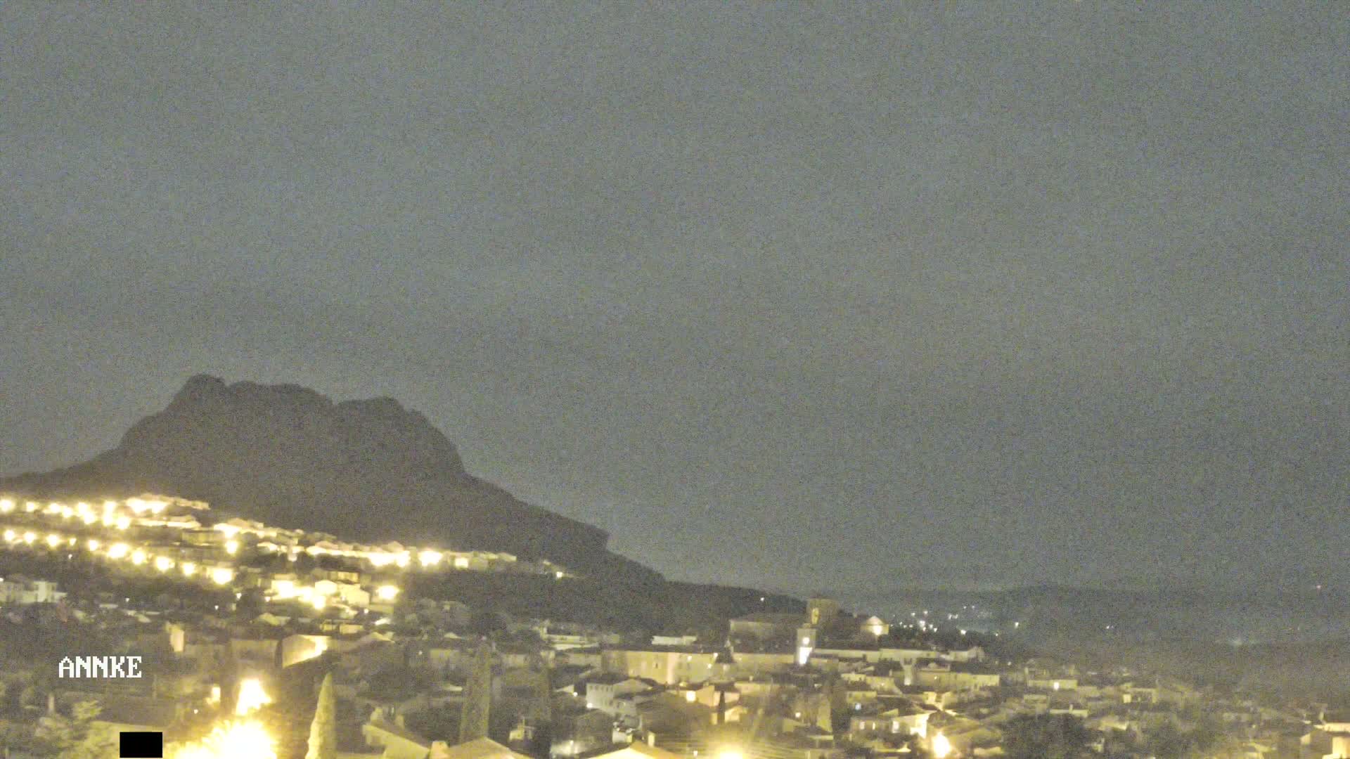 A nighttime view of a town illuminated by lights, nestled beneath a dark, silhouetted mountain under a cloudy sky.
