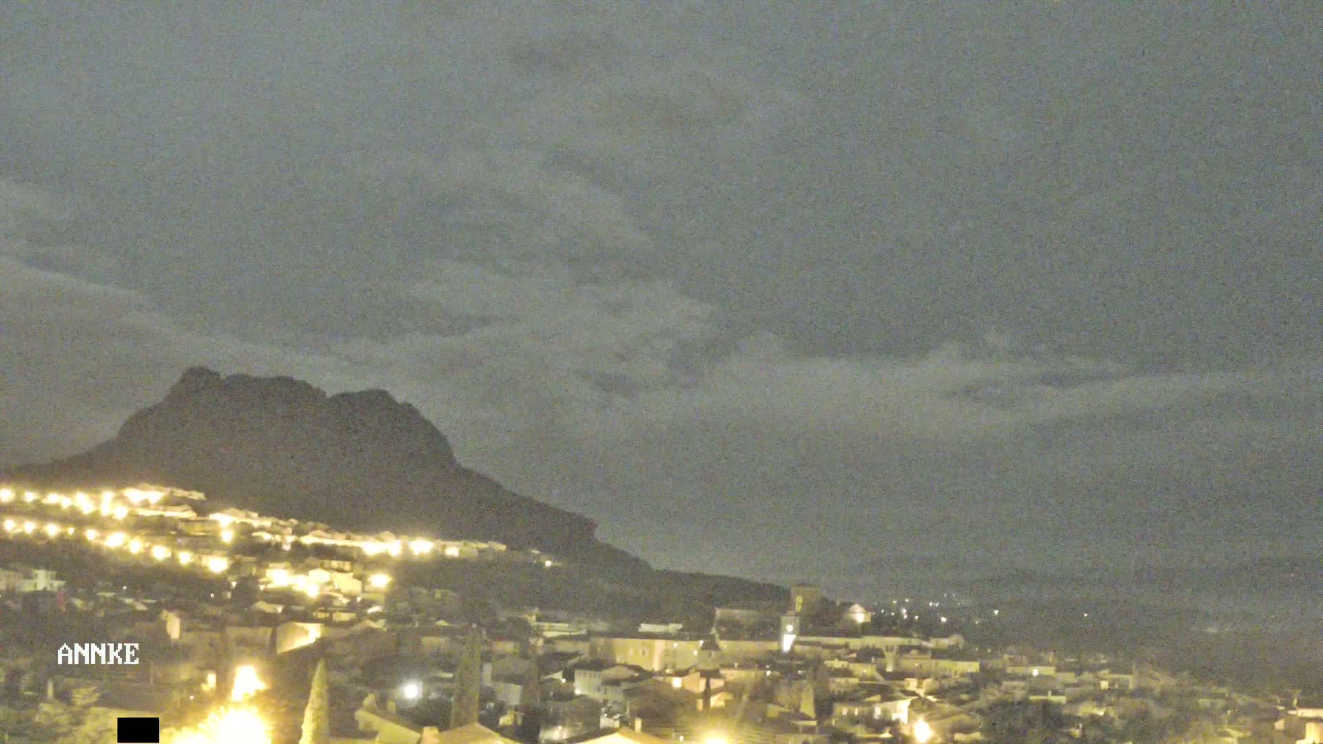 A nighttime view of a town nestled at the base of a dark mountain under a cloudy sky.