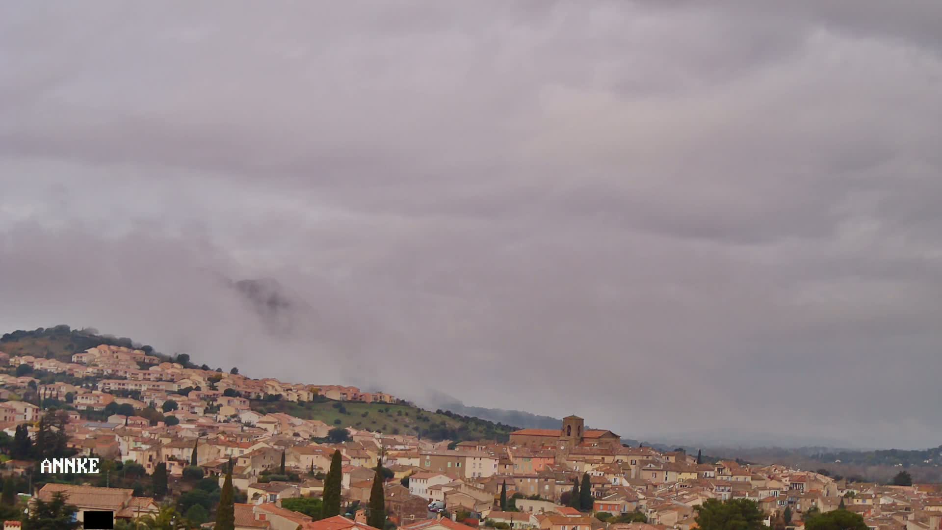 A hillside town with many tan and brown buildings is covered in a layer of low-hanging clouds under an overcast sky.