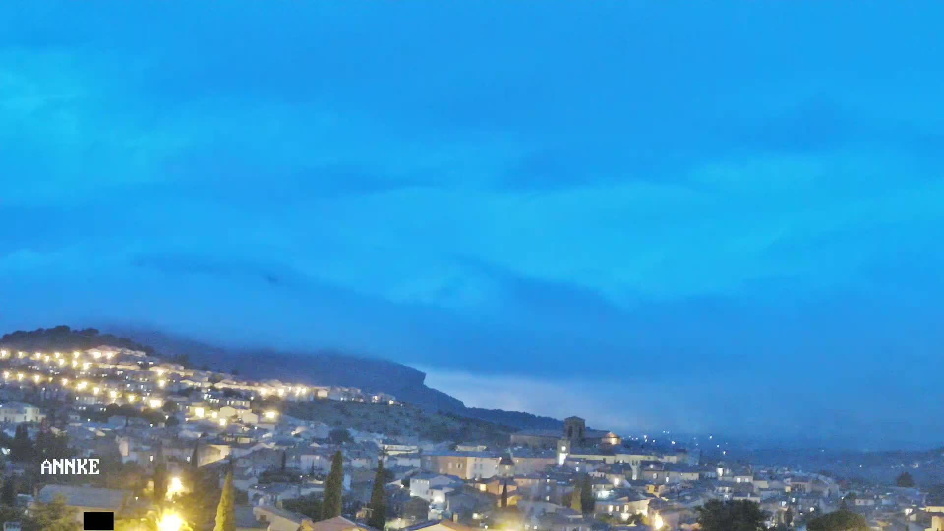 A hilltop town is illuminated at night under a mostly clear, blue sky with a low layer of clouds or fog clinging to the hills.