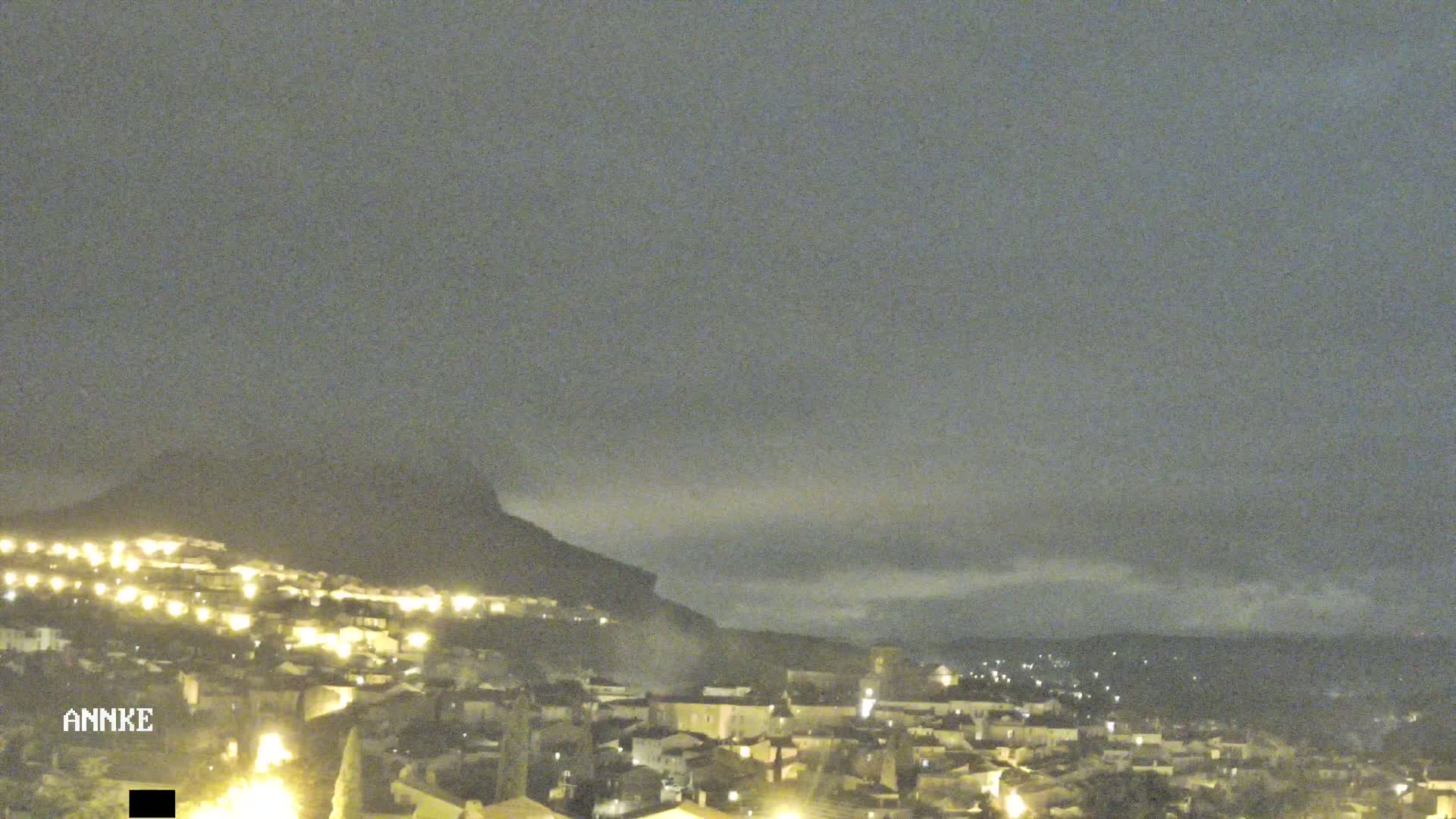 A nighttime view of a town nestled at the base of a dark mountain under a cloudy sky.