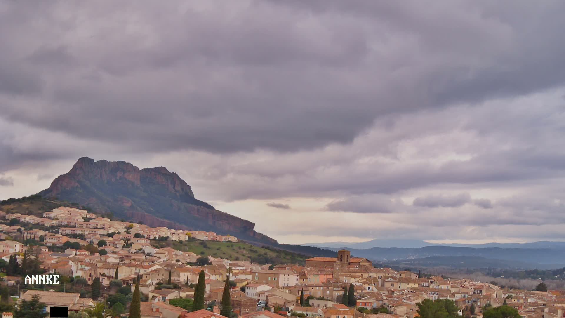 A sprawling village of tan and brown buildings nestles against a steep, dark-colored mountain under a cloudy sky.