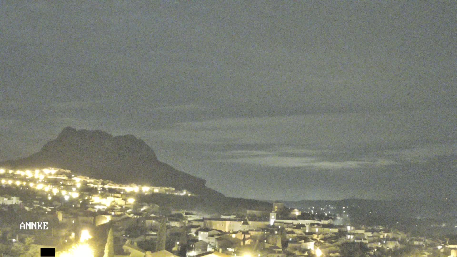 A nighttime view of a town illuminated by lights nestled below a dark mountain under a cloudy sky.