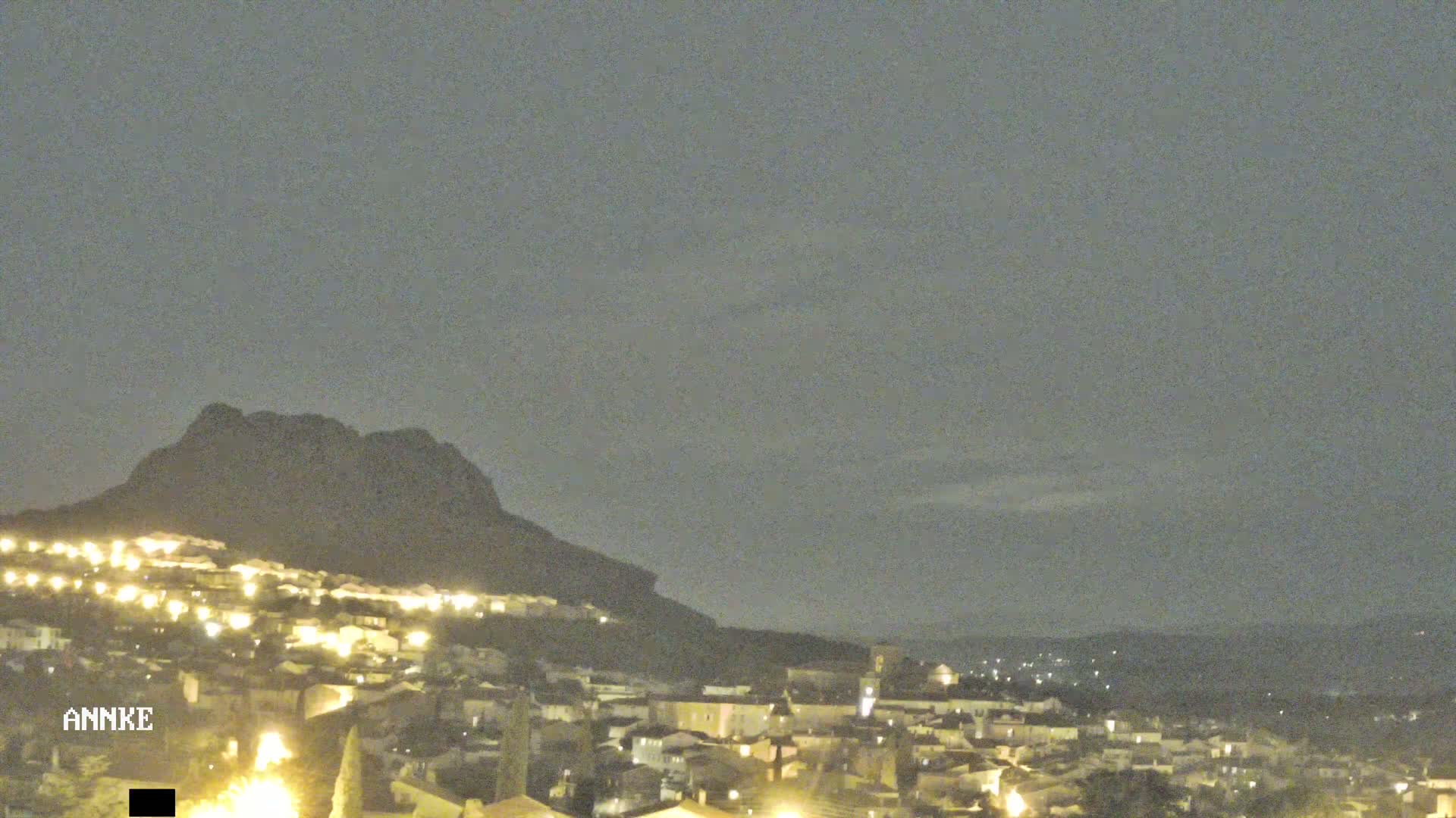 A nighttime view of a town nestled at the base of a large, dark mountain under a hazy, overcast sky.