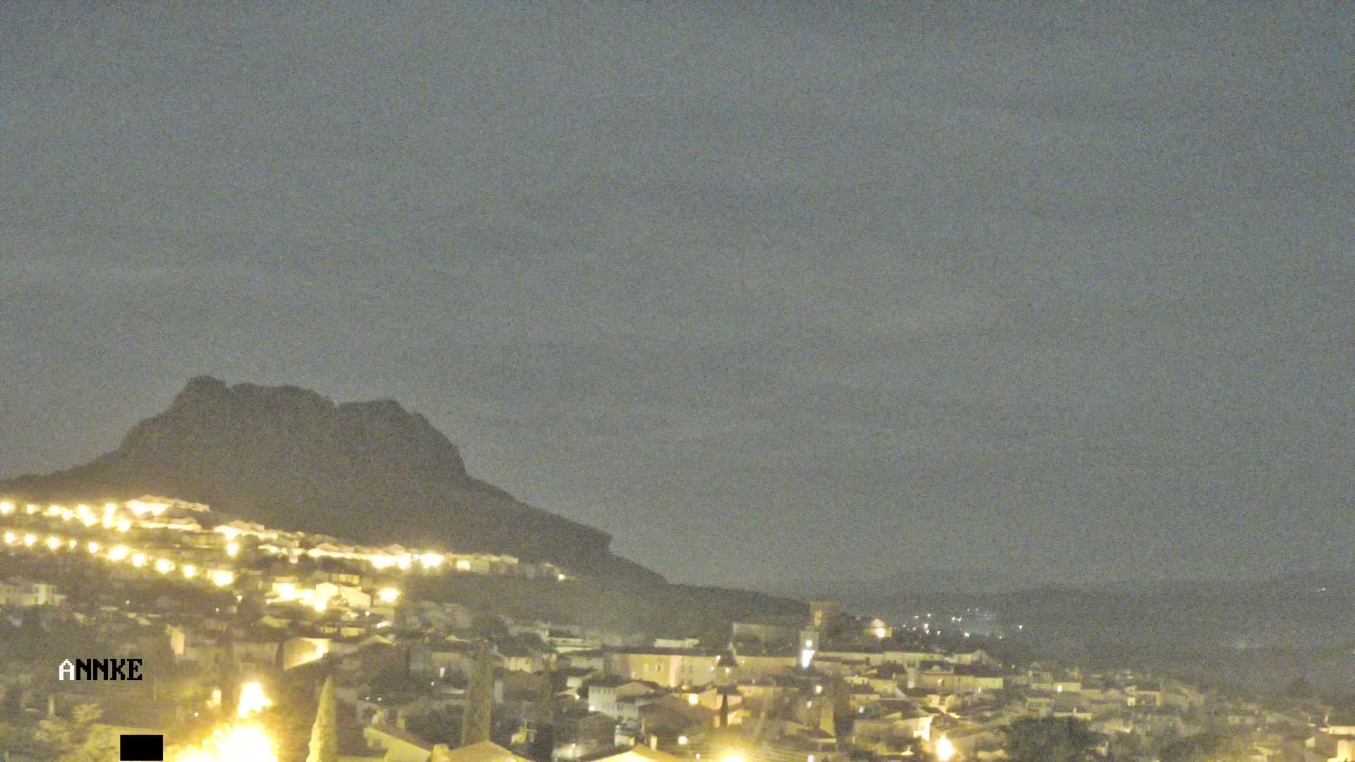 A nighttime view of a town nestled at the base of a dark mountain under a cloudy sky.