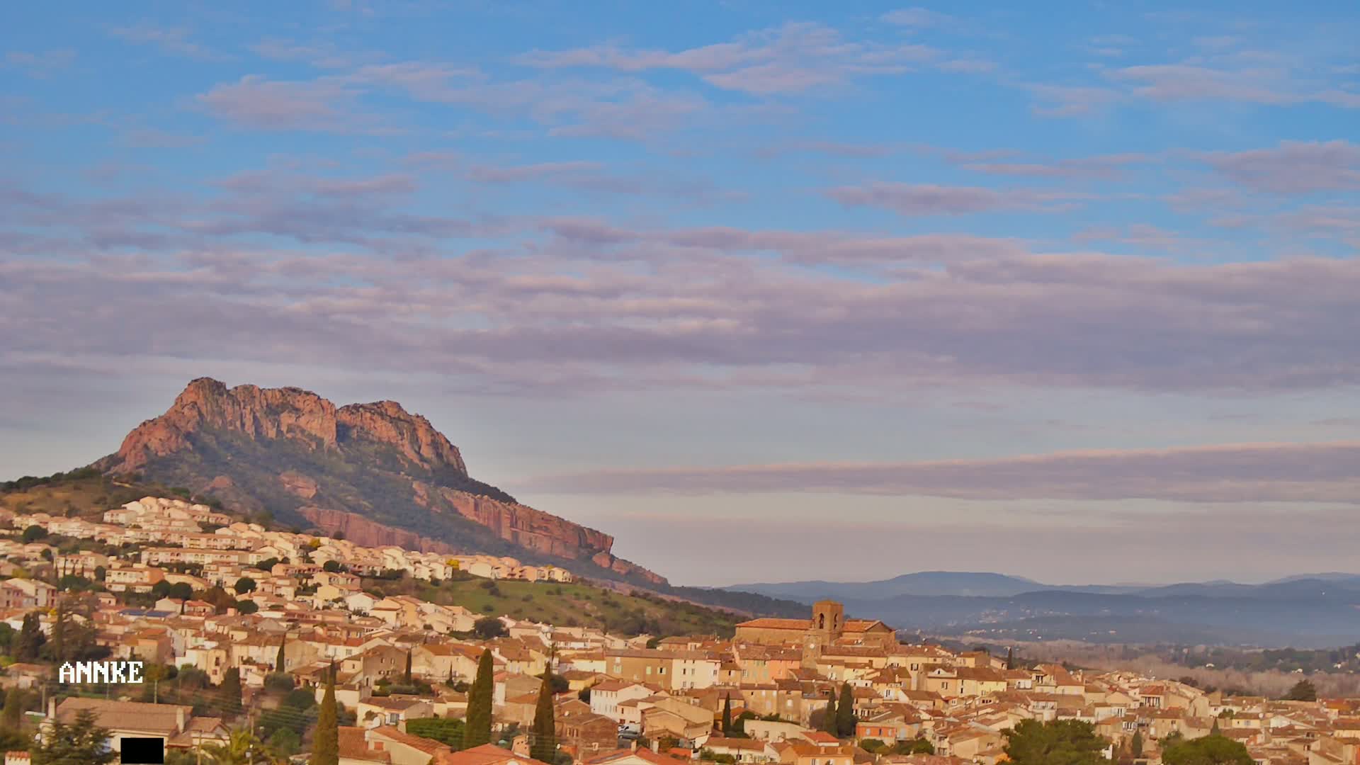 A town of tan-colored buildings nestled against a reddish-brown hill under a mostly clear sky with faint purple clouds.