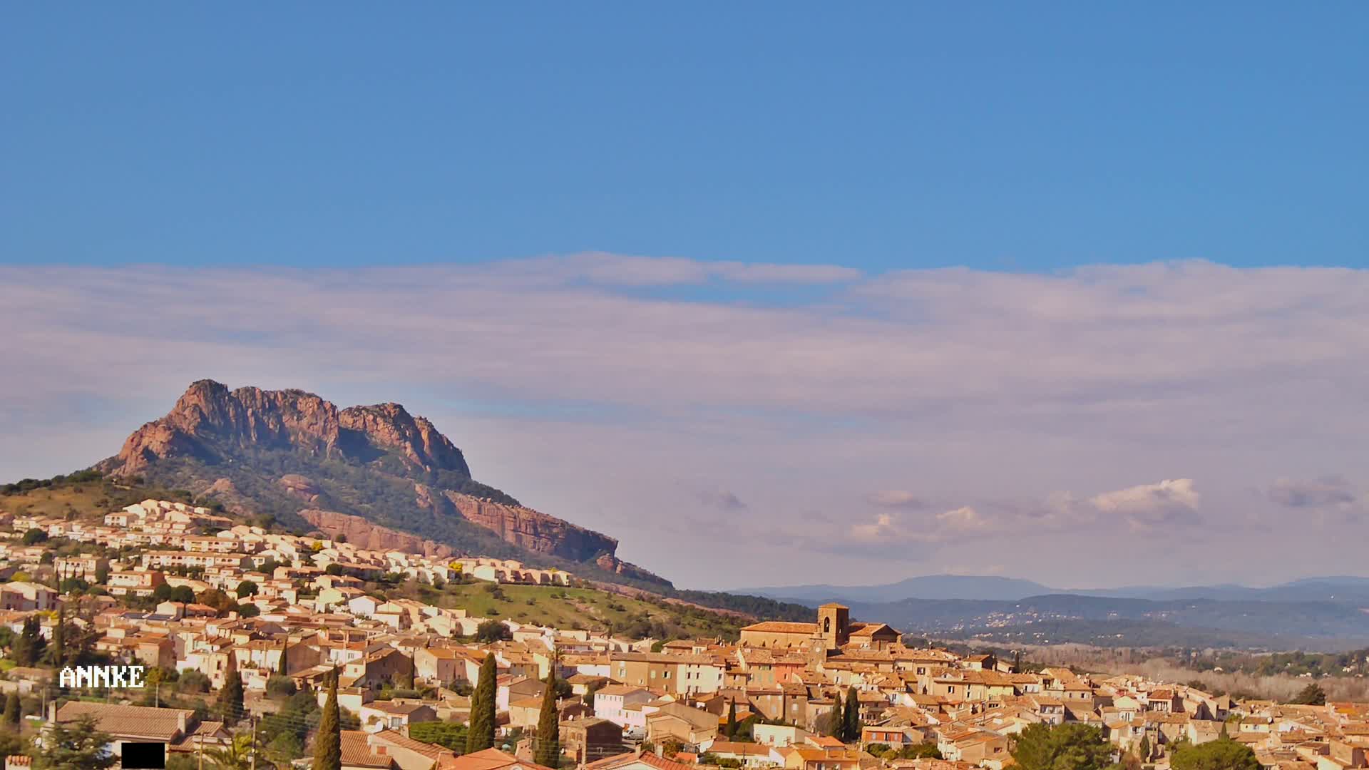 A sprawling town of tan buildings nestled on a hillside beneath a large, reddish-brown mountain under a mostly clear, light blue sky with a few wispy clouds.