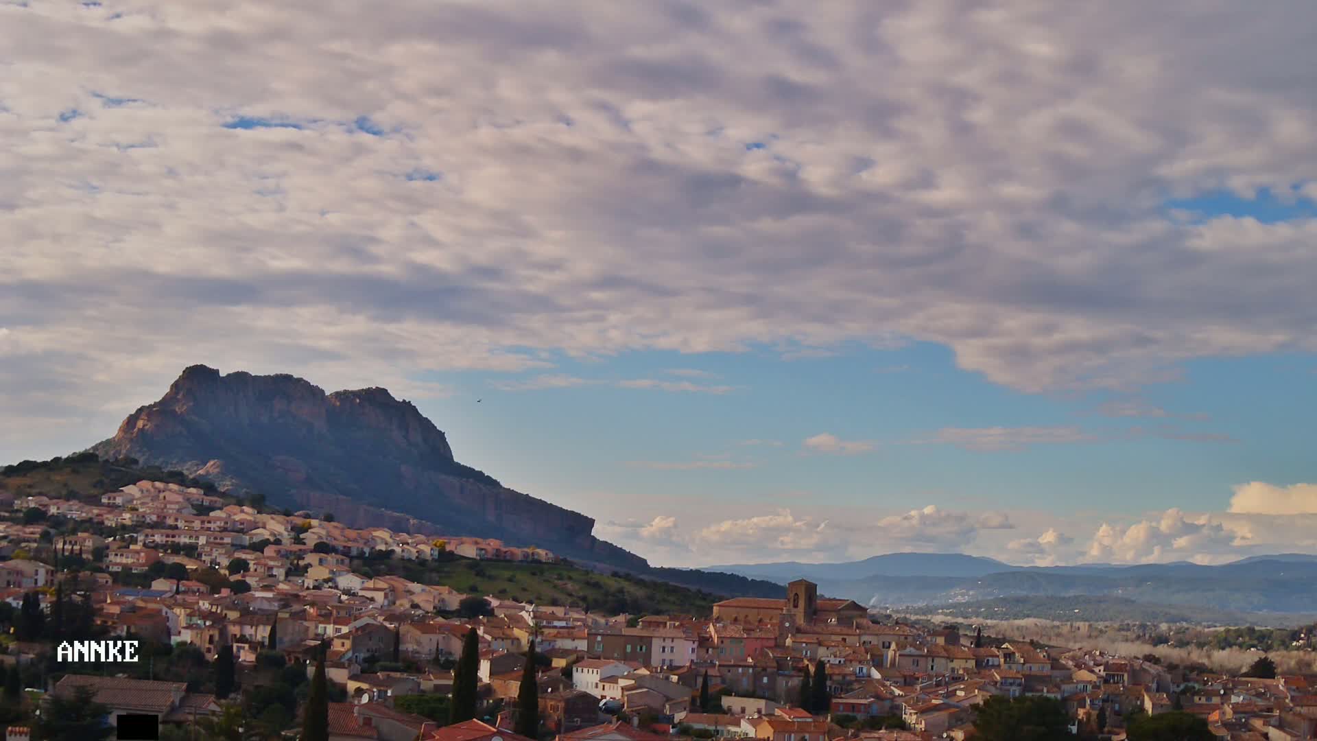 A large hill covered in a small town sits under a mostly cloudy sky with distant mountains visible on the horizon.