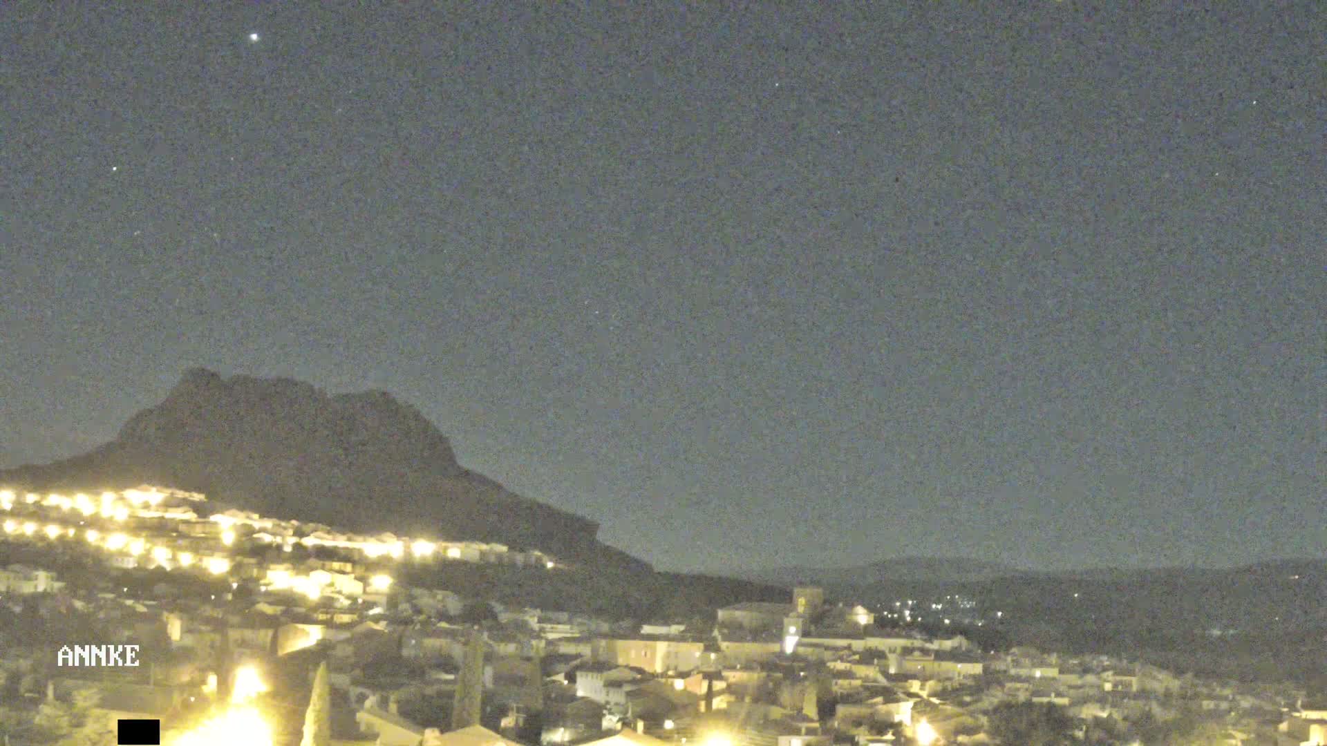 A nighttime view shows a town illuminated by lights, nestled at the base of a large, dark mountain under a clear, starlit sky.