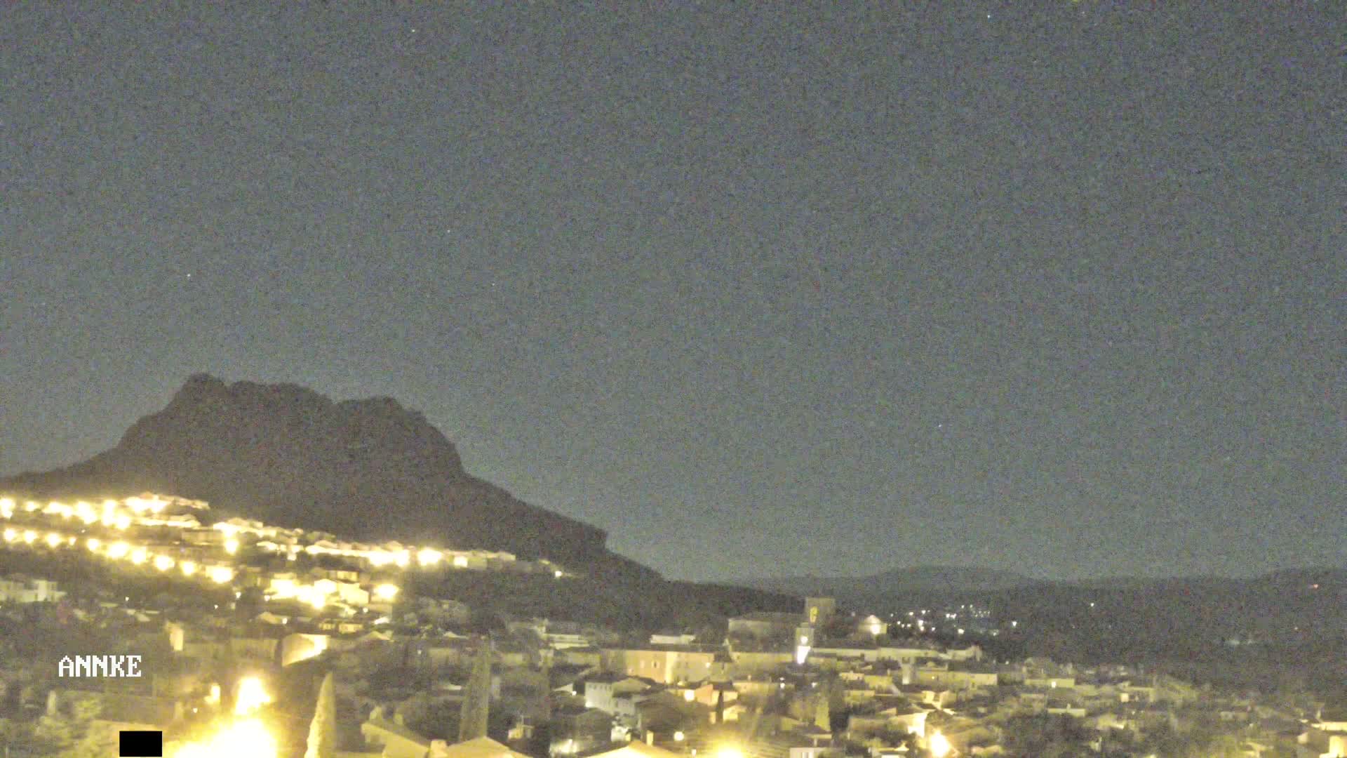 A nighttime view of a town nestled at the base of a dark mountain under a clear, star-studded sky.