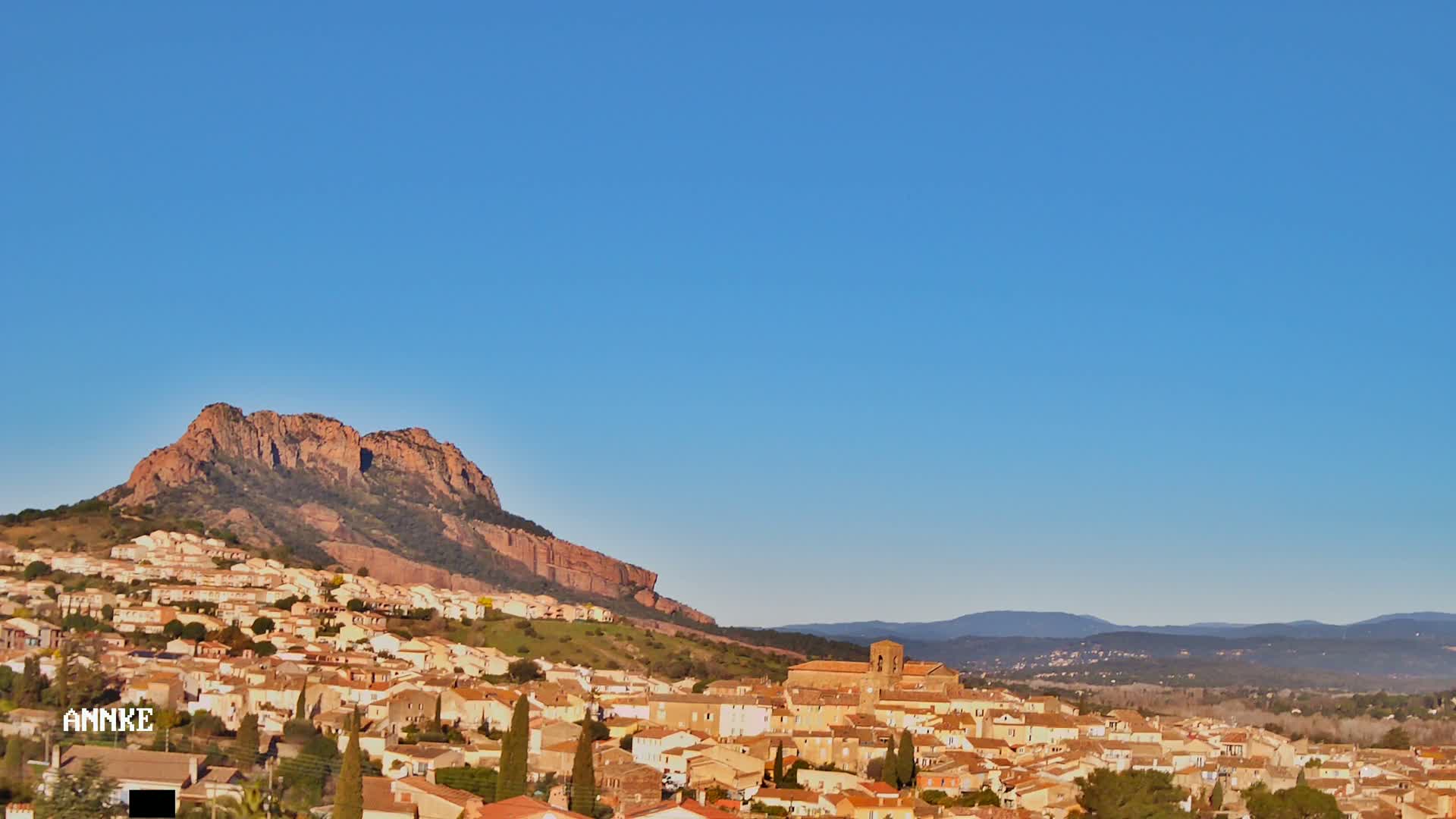 A large town of tan buildings nestled against a reddish-brown mountain under a clear blue sky.