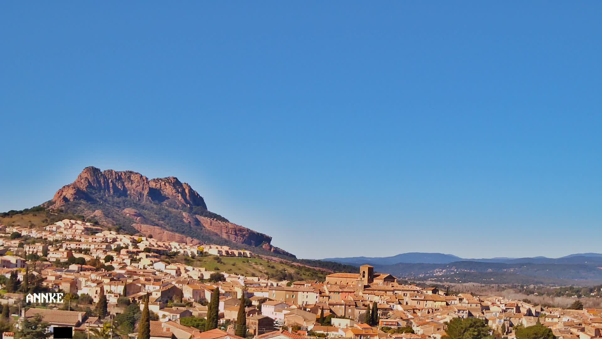 A sprawling town of tan and brown buildings nestled against a large, reddish-brown mountain under a clear blue sky.