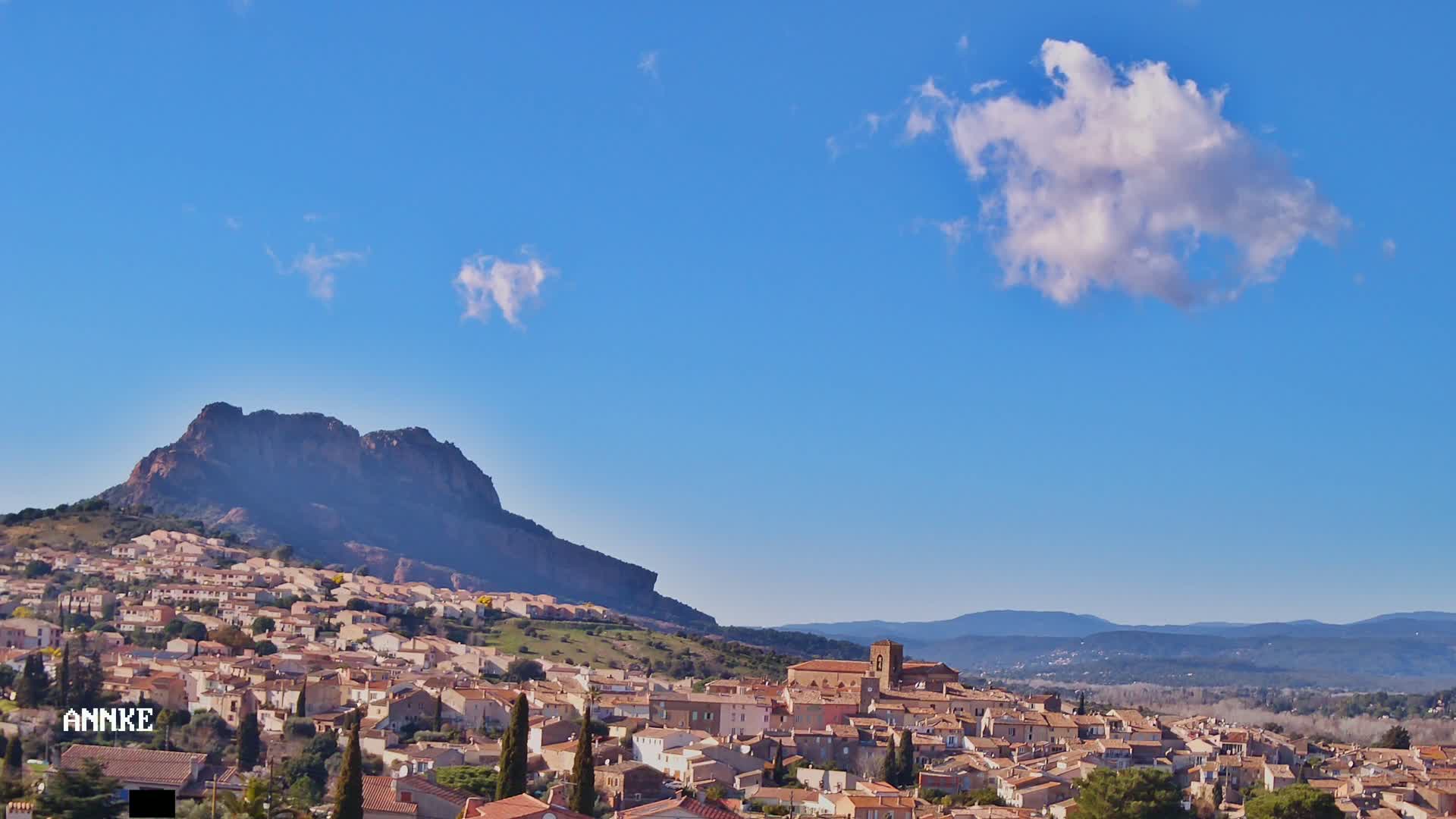 A sprawling town of tan and brown buildings nestles at the base of a large, dark mountain under a clear blue sky with a few fluffy white clouds.