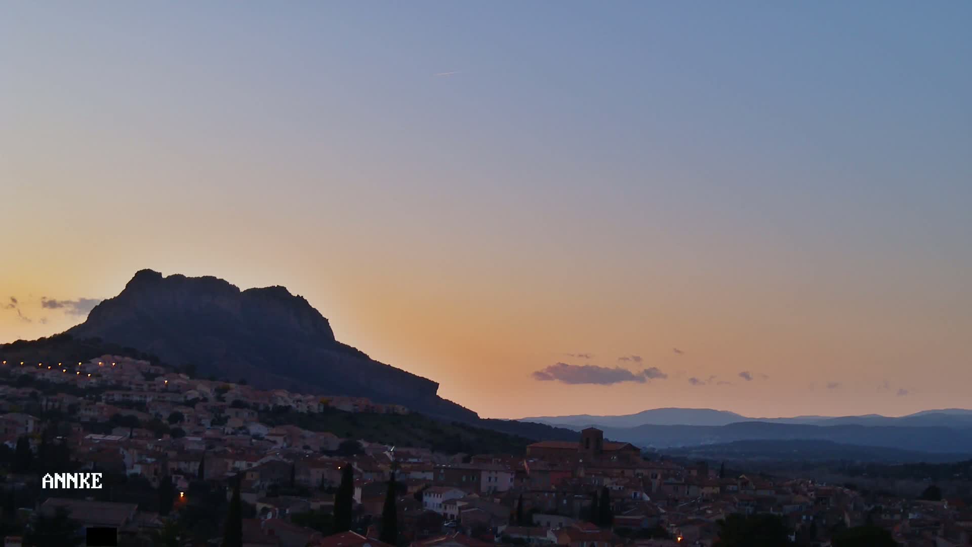 A town nestled at the base of a large, dark mountain, silhouetted against a pastel sunset sky.