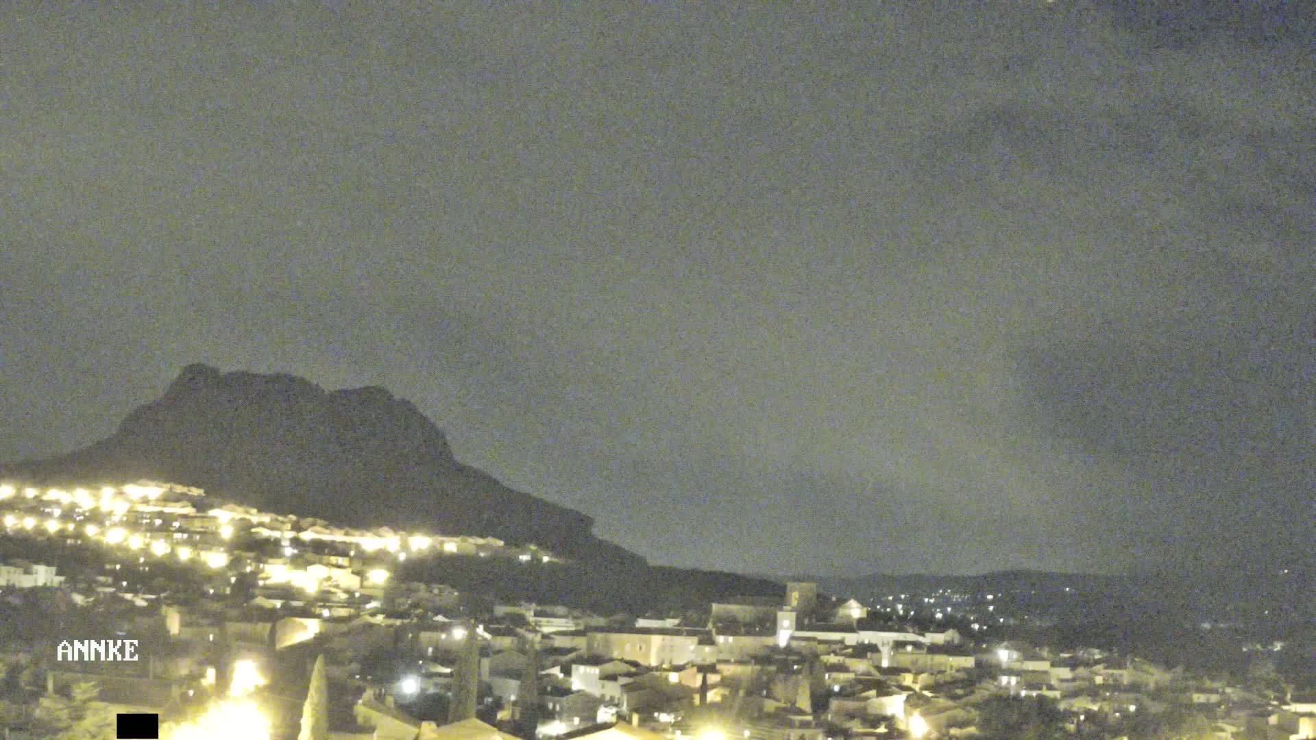 A nighttime view of a village nestled at the base of a dark mountain under a cloudy sky.