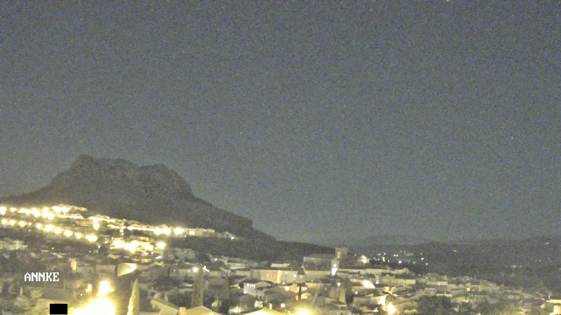 A nighttime view of a town illuminated by lights, nestled at the base of a dark mountain under a clear, dark sky.