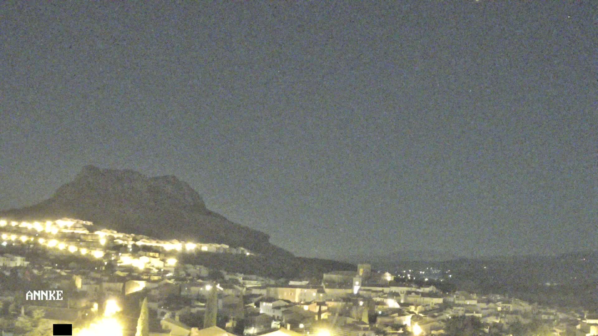 A nighttime view of a town nestled at the base of a dark mountain under a clear, dark sky.