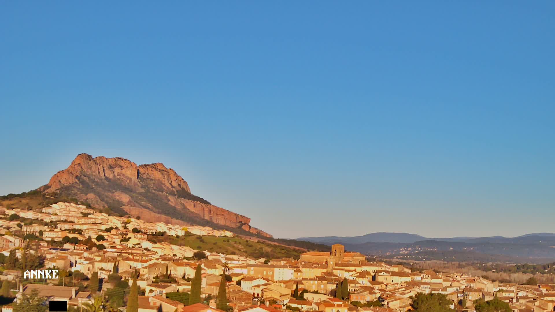 A small town nestled against a large, reddish-brown mountain under a clear blue sky.