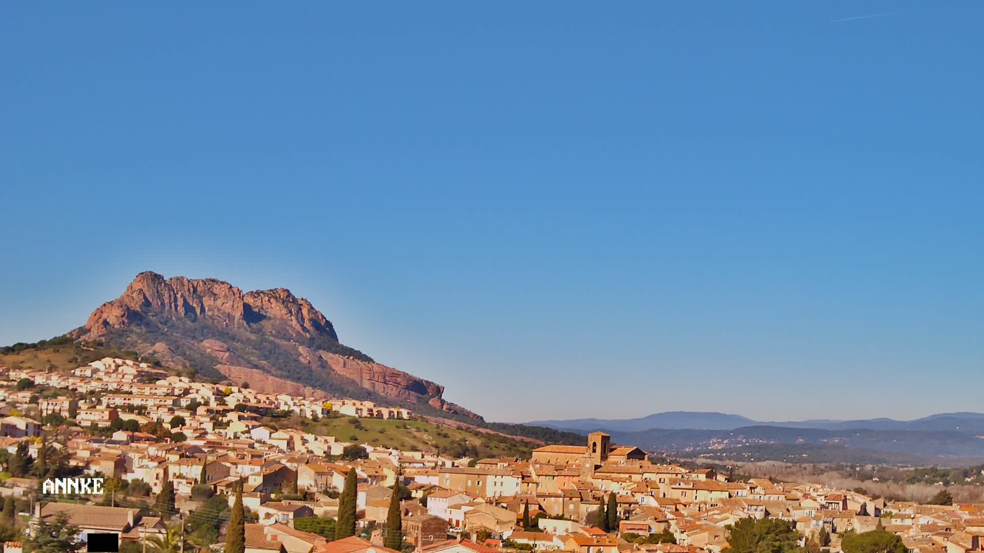 A town of tan and brown buildings nestled against a rocky, reddish-brown mountain under a clear blue sky.