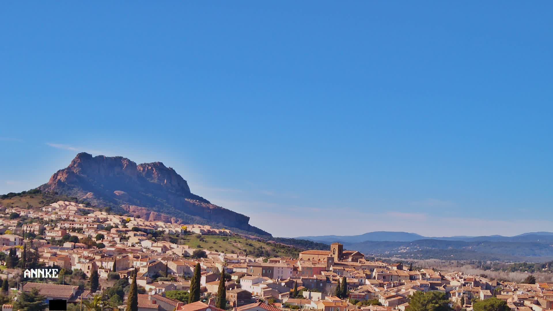 A small town with tan buildings nestles at the base of a large, rocky mountain under a clear blue sky.