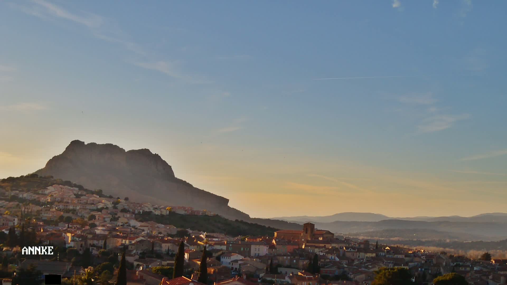 A small town nestled at the base of a large, imposing mountain under a clear, pale sunset sky.