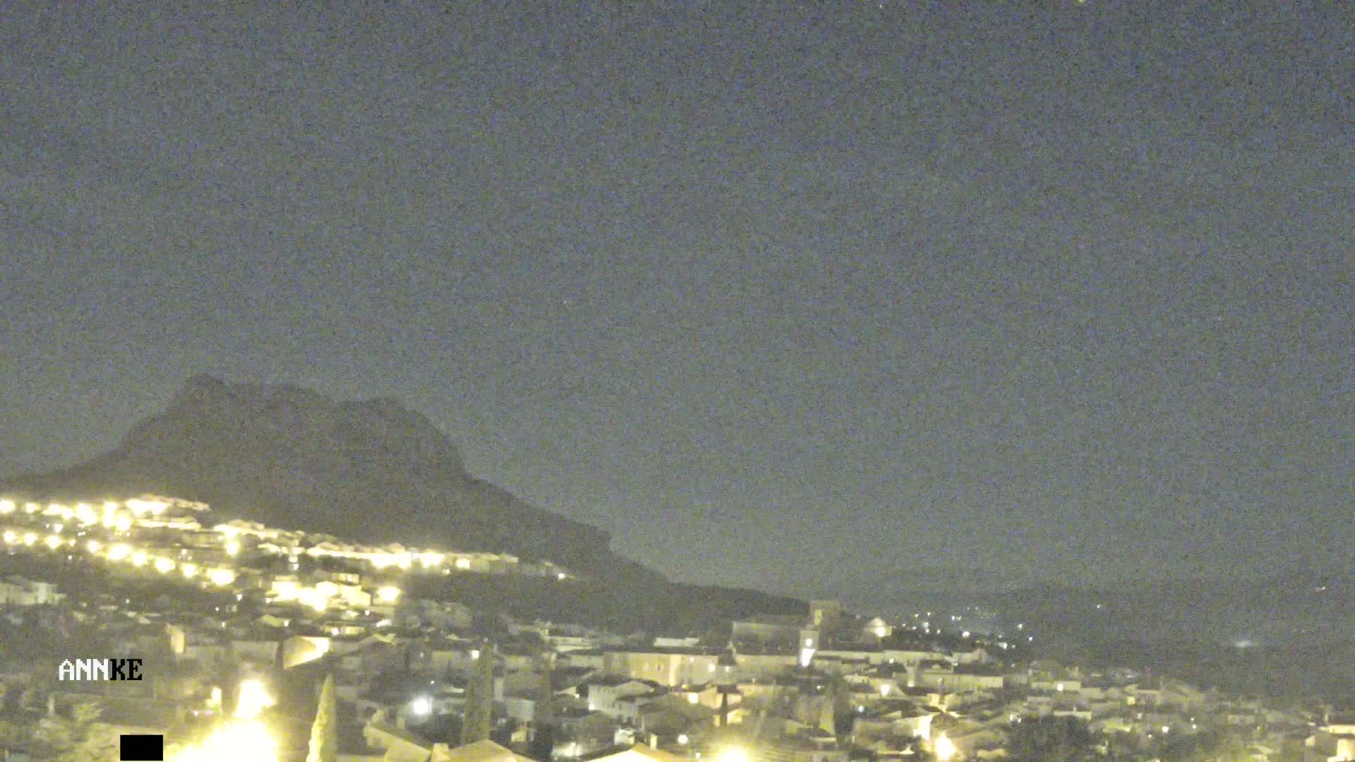 A nighttime long shot shows a town nestled at the base of a hill under a clear, dark sky.