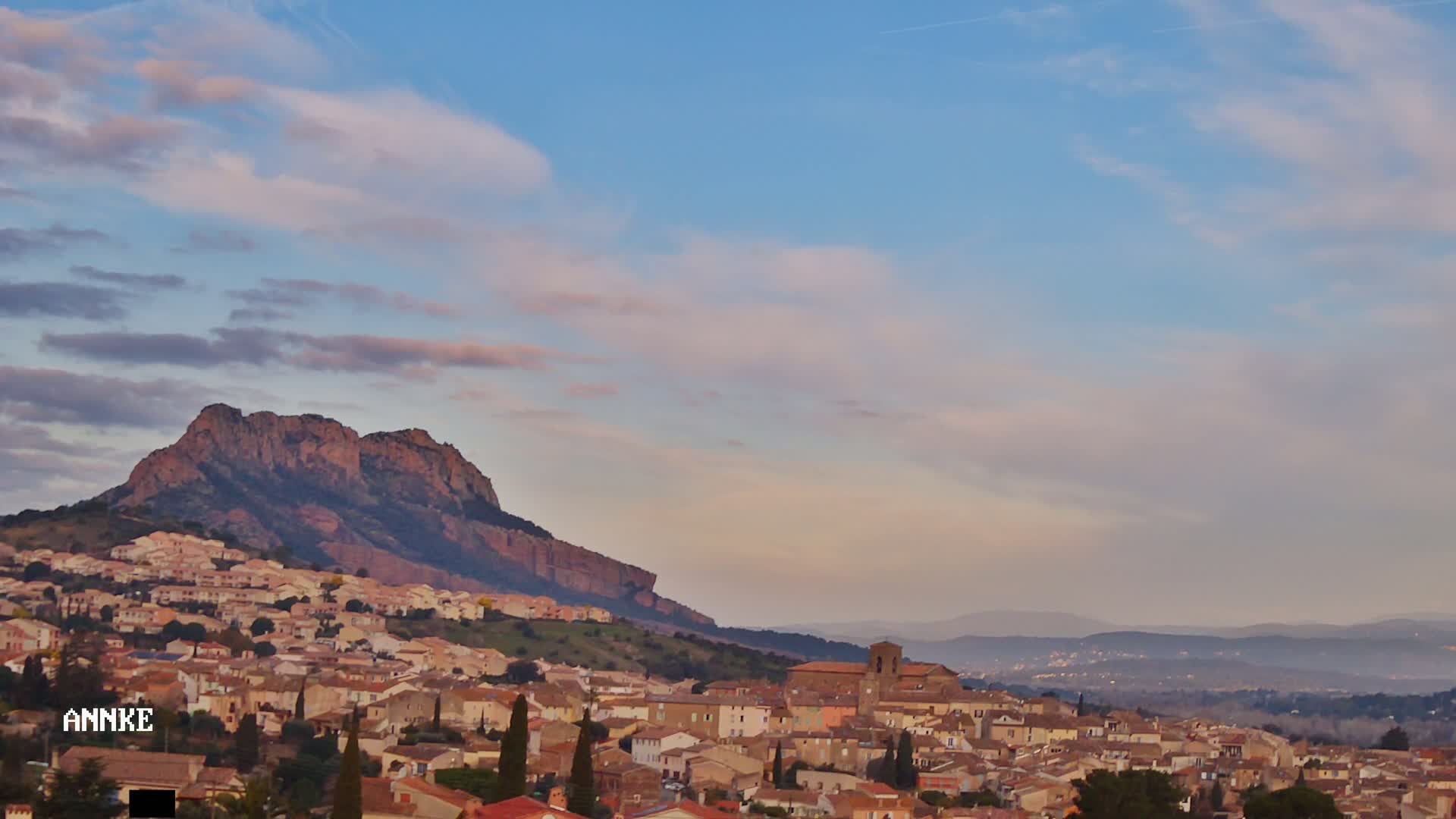 A hilly town with tan buildings nestles beneath a large, reddish-brown mountain under a mostly clear, pale blue sky with thin, wispy clouds.