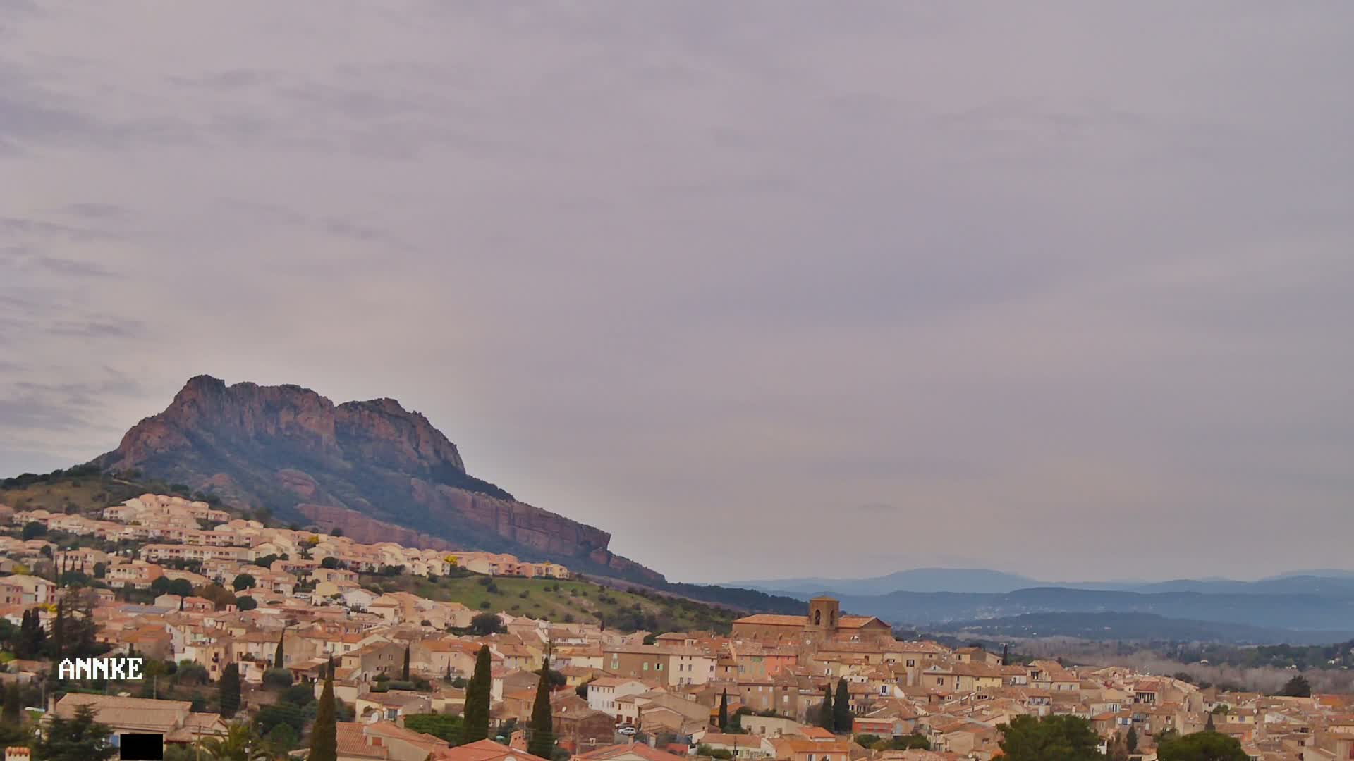 A village of tan and brown buildings sprawls across a hillside beneath a large, dark mountain under a cloudy sky.