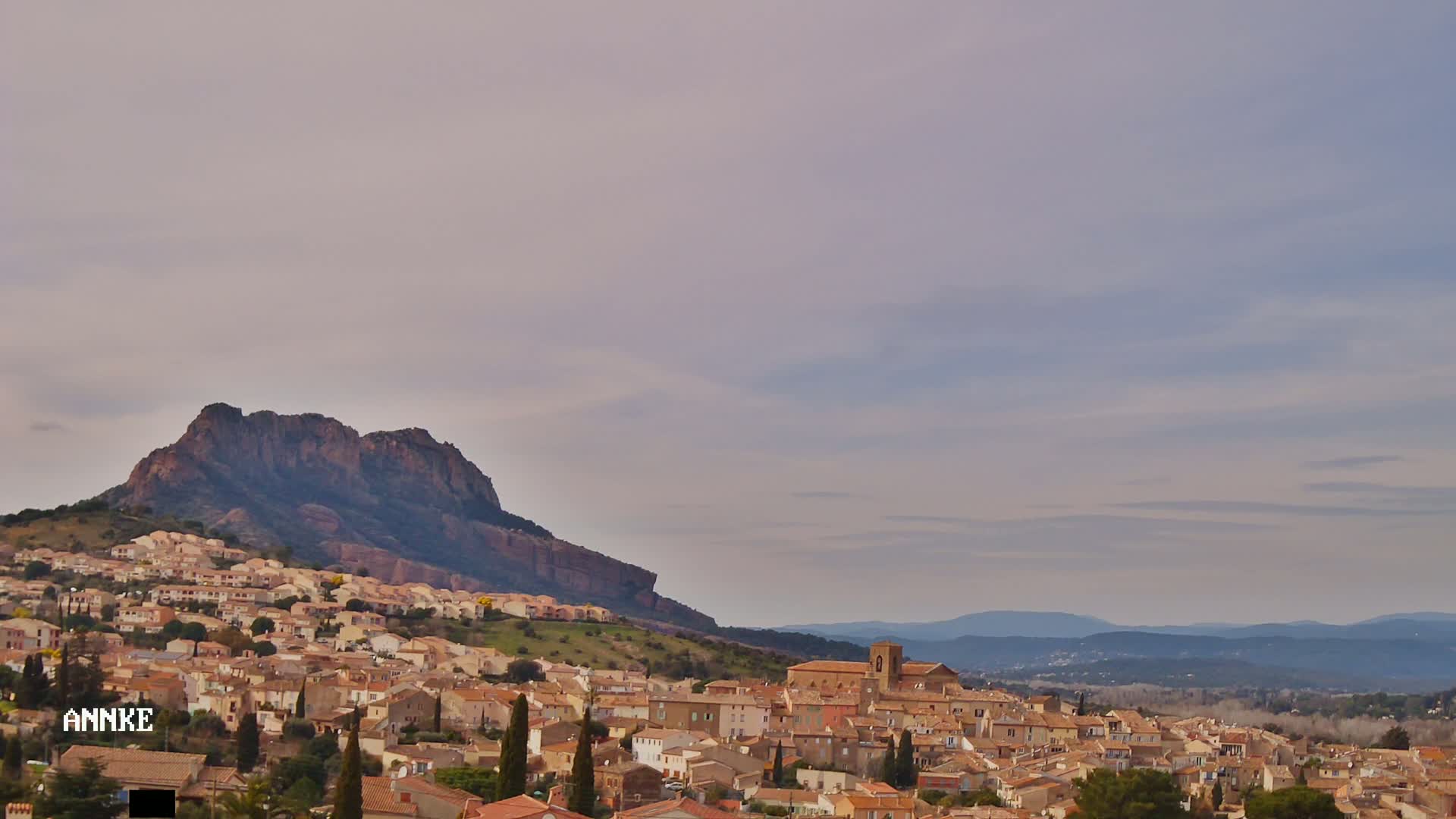A large, tan-colored town nestled against a rocky, dark-brown mountain under a pale, mostly cloudy sky.