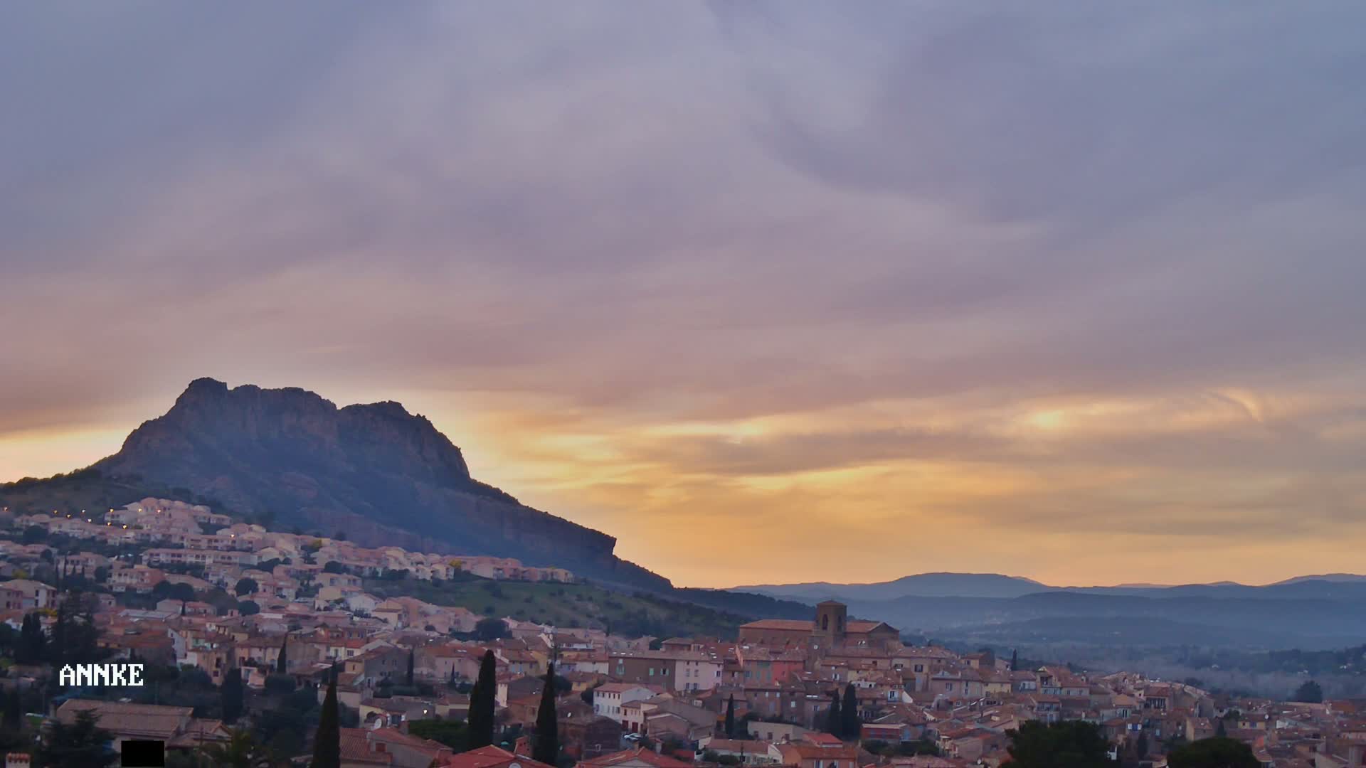 A hilltop town with terracotta roofs nestles beneath a large, dark mountain against a pastel sunset sky.
