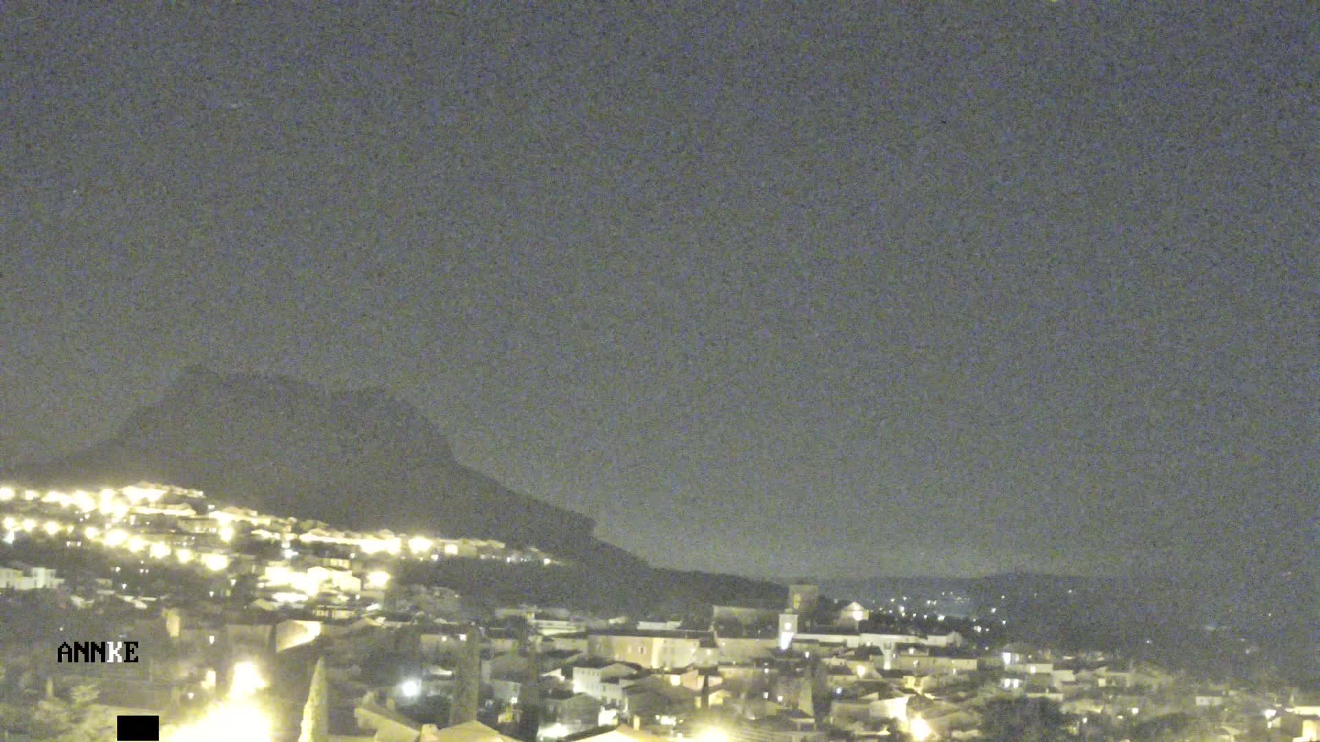 A nighttime view of a town nestled against a dark mountain, under a clear, dark sky.