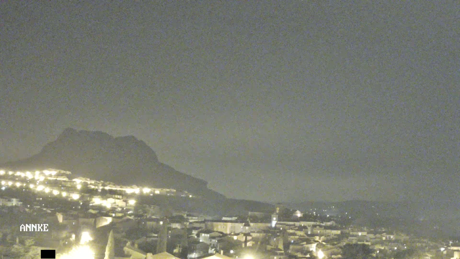 A nighttime view of a town nestled at the base of a dark mountain under a cloudy sky.