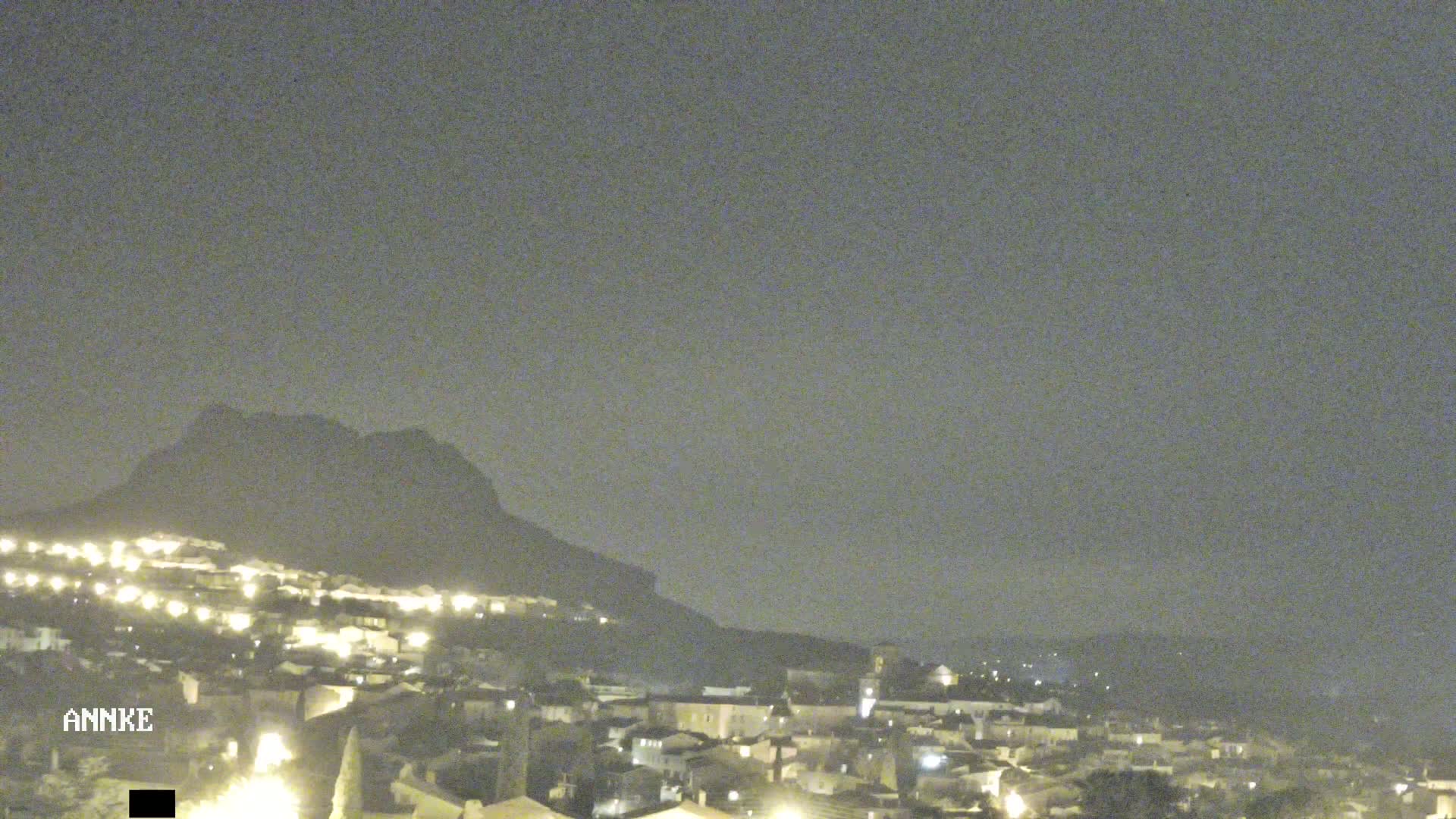 A nighttime view of a town nestled at the base of a dark mountain under a hazy sky.