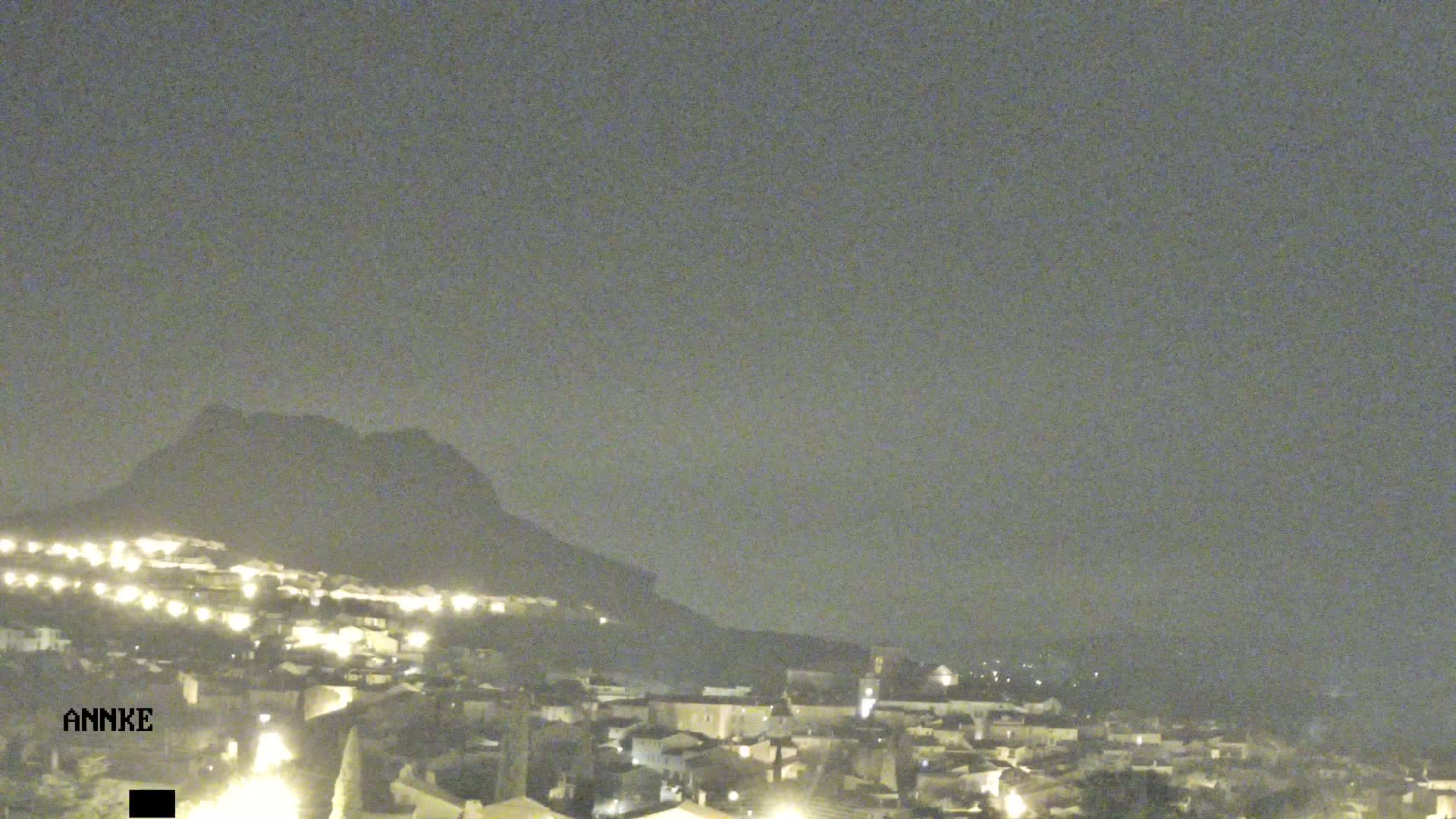 A nighttime view of a town nestled at the base of a hill under a hazy sky.