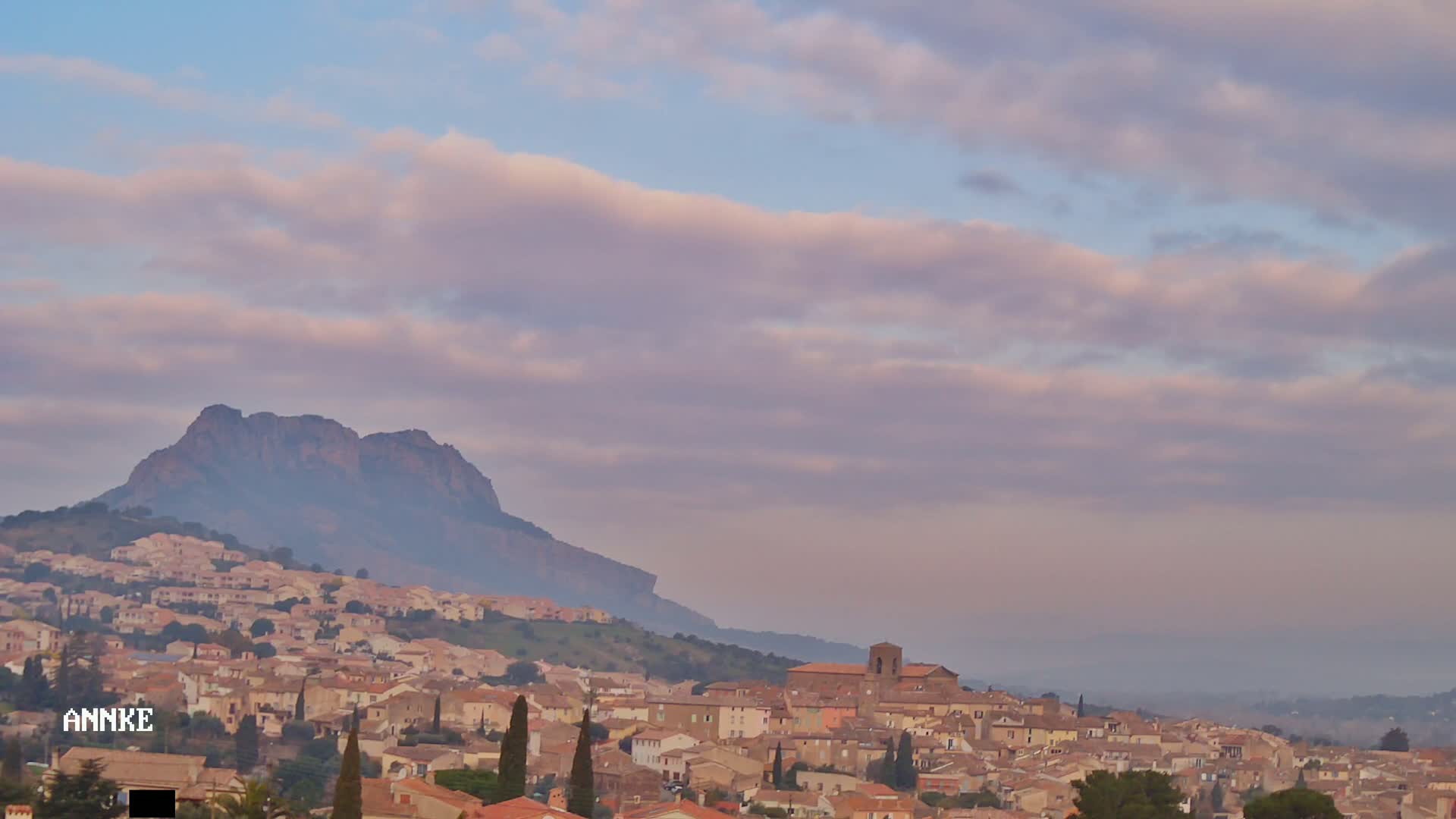 A sprawling town of tan and brown buildings nestles at the foot of a large, dark mountain under a soft, pale pink and blue sky.