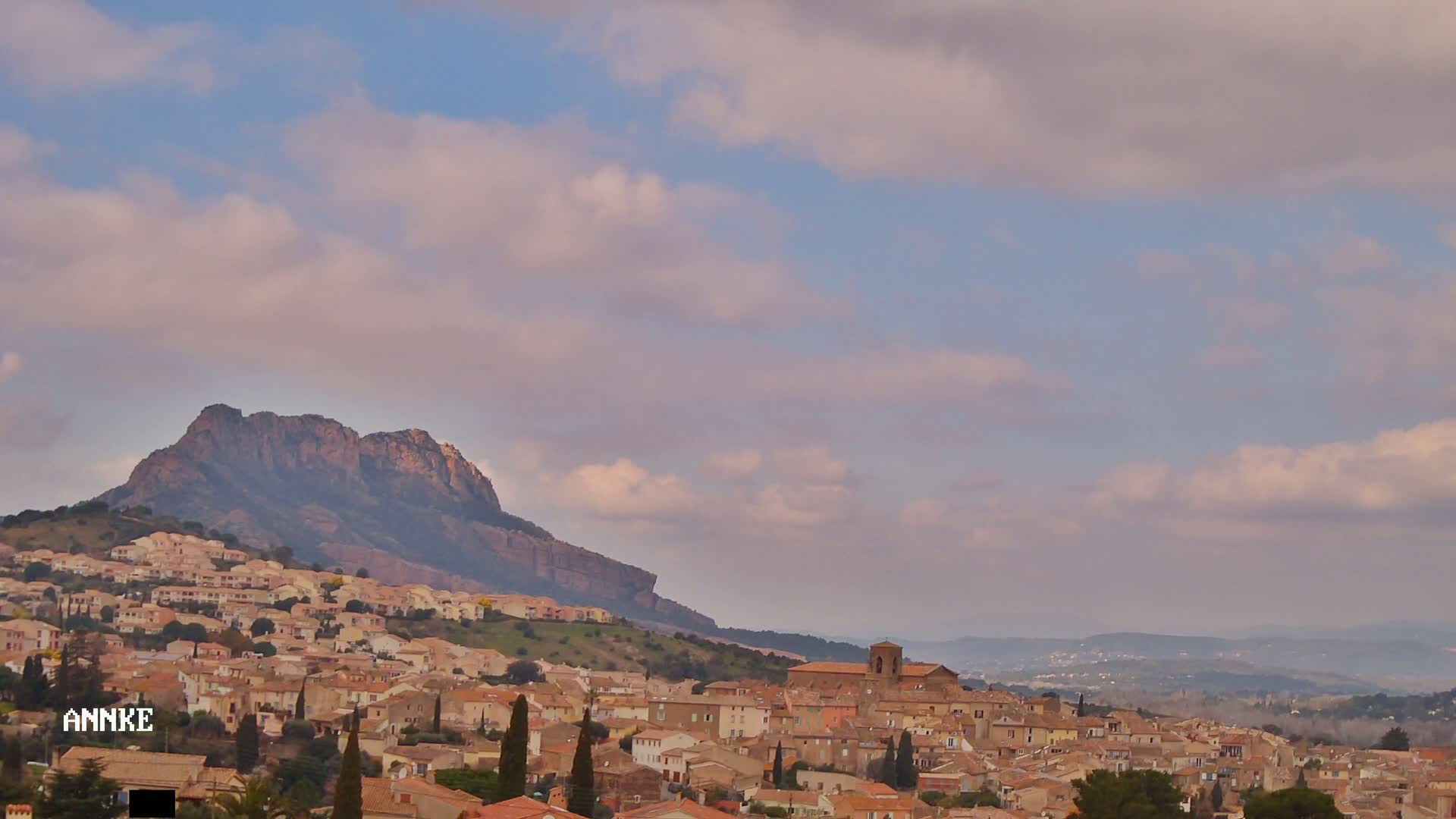 A sprawling hillside town with light-brown buildings nestles beneath a large, dark mountain under a partly cloudy sky.