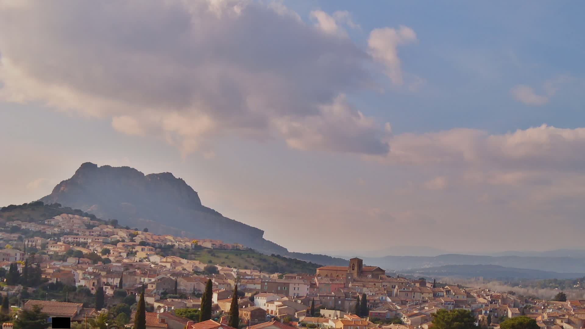 A quaint town nestled at the base of a large mountain under a partly cloudy sky.